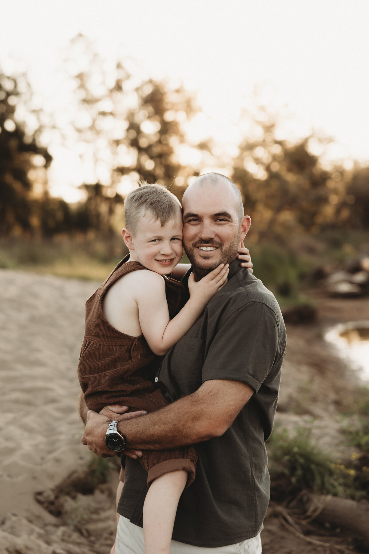A dad holds a young child on a sandy path with trees and soft sunlight in the background. Both are smiling and wearing casual clothing, perfectly capturing the essence of South Coast family photography.