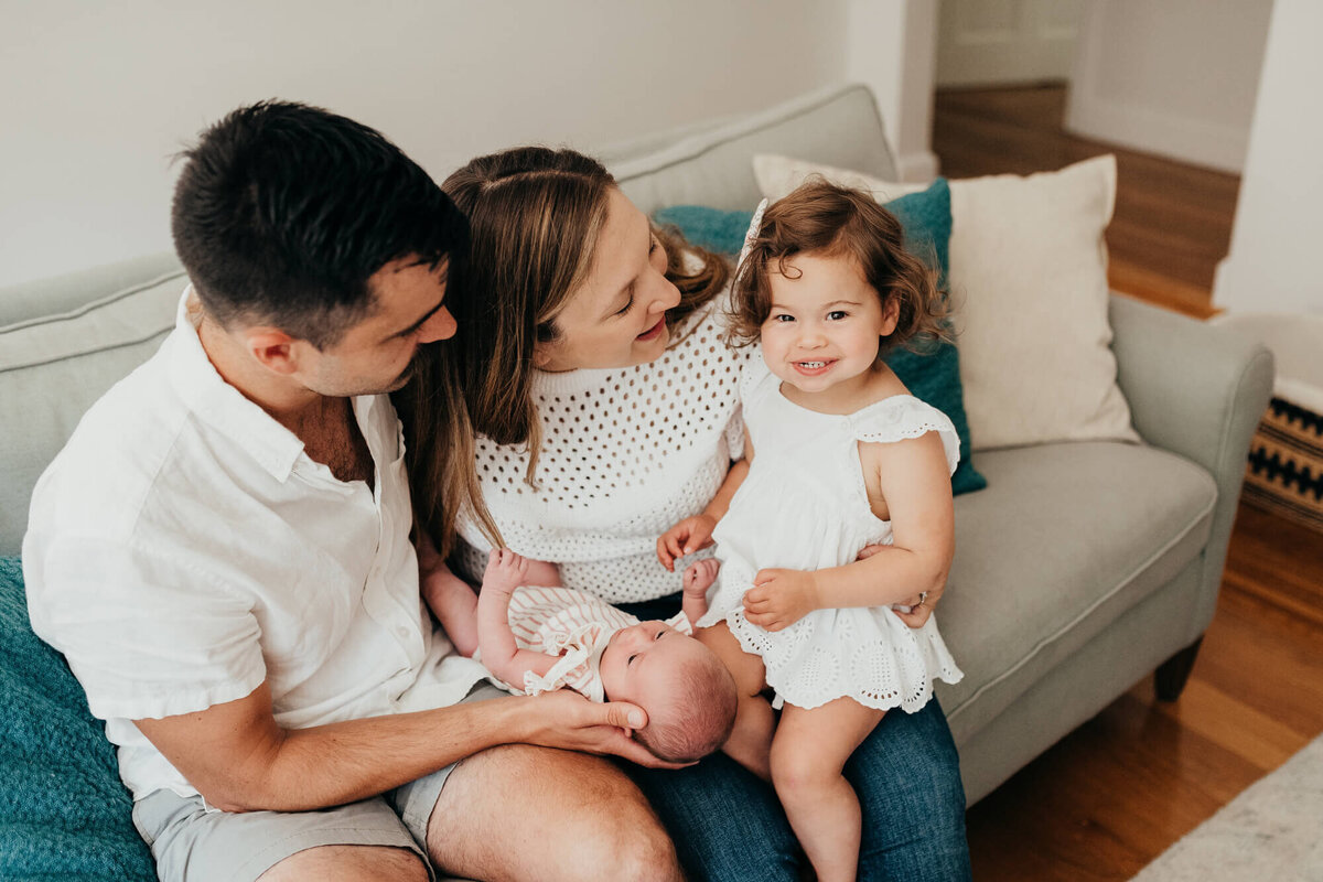 A family with two young daughters sit together on their couch in a bright living room