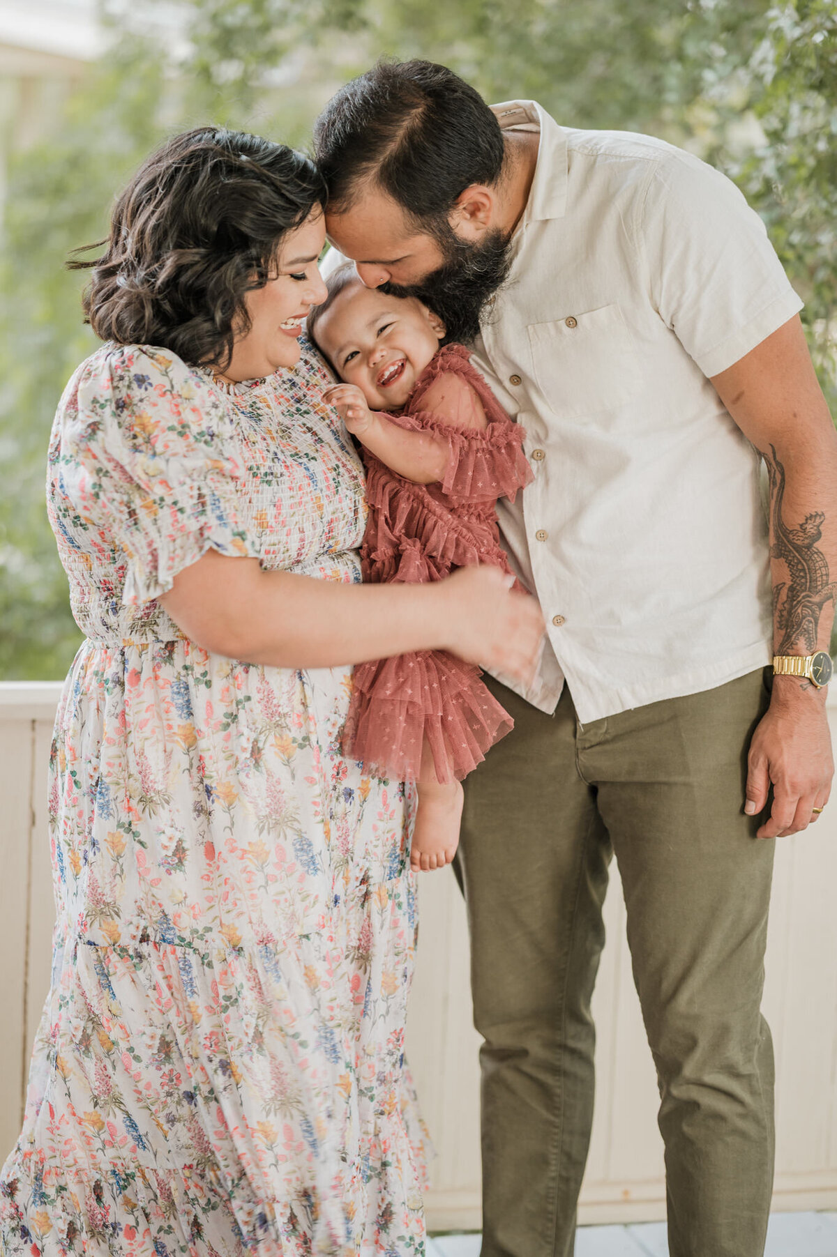 Mom and dad smile and kiss their laughing daughter during family pictures.
