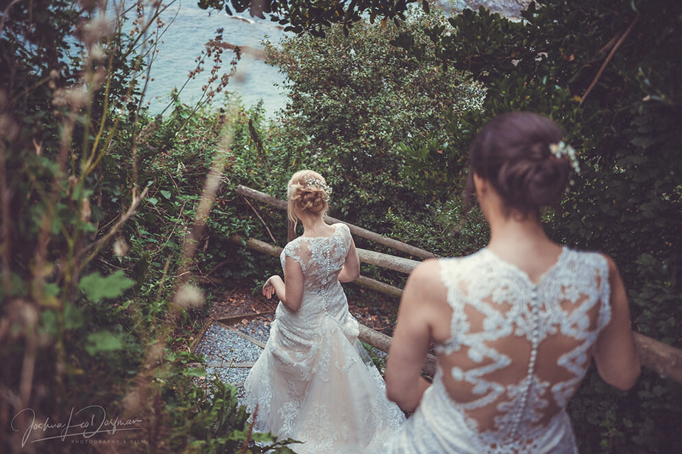 Two women in elegant, lace wedding dresses walk down a leafy, outdoor staircase towards a body of water. The women are seen from behind, highlighting the intricate lace details on the backs of their dresses. Nature surrounds them with green foliage on both sides.