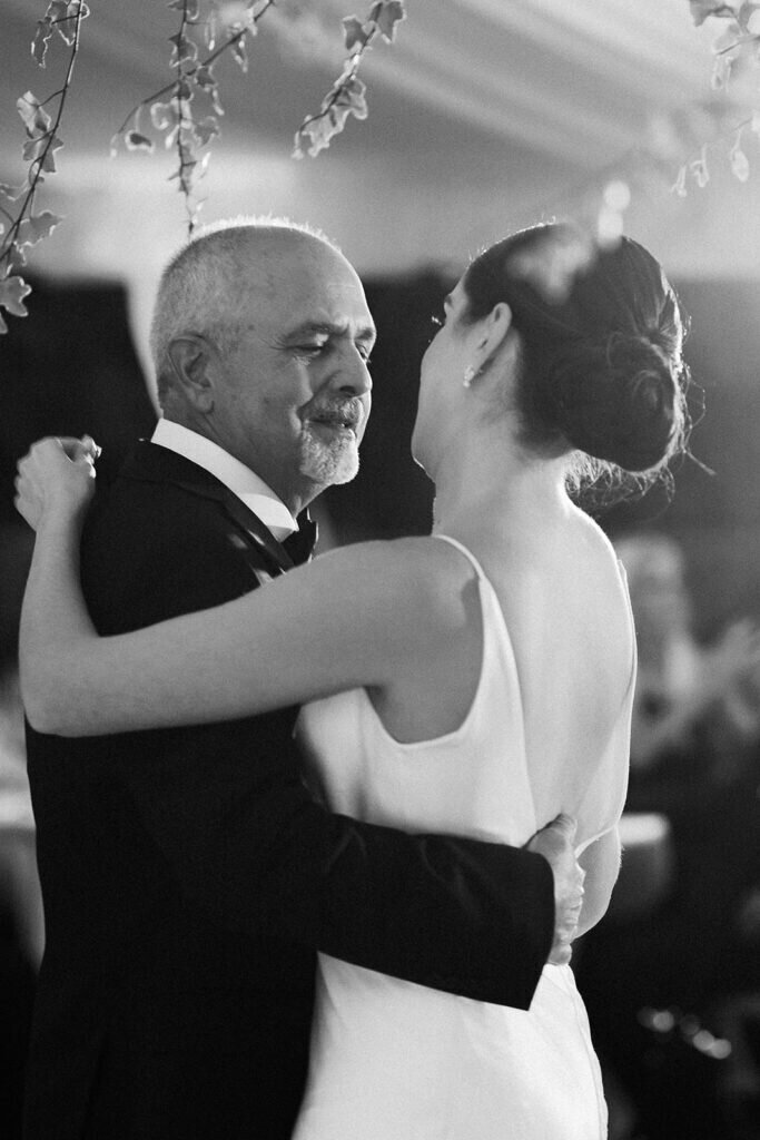 A heartfelt black-and-white moment as the bride shares an emotional dance with her father, surrounded by warm wedding reception lighting and lush greenery. Captured by Claudia Amalia Photography, a Florida Keys wedding photographer.