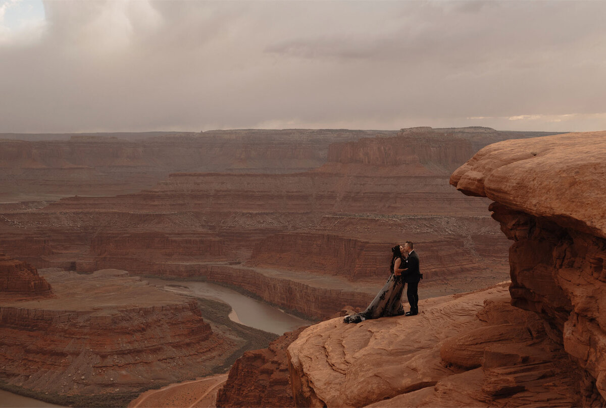 elopement couple kisses with the grandeur of the Dead Horse Point State Park in Moab, Utah scenery in the background on a moody day