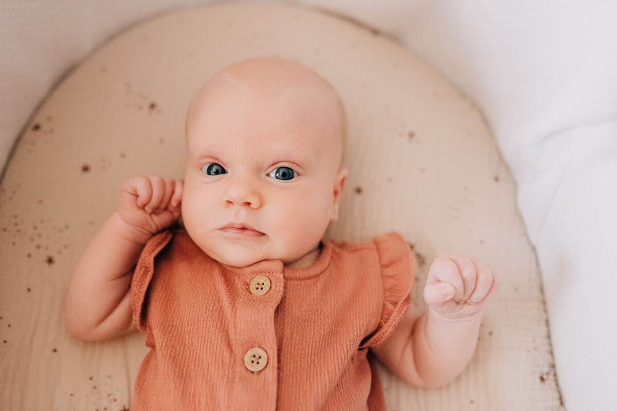 A newborn baby girl looks at the camera with bright eyes wearing a rose colored outfit and laying in a cradle