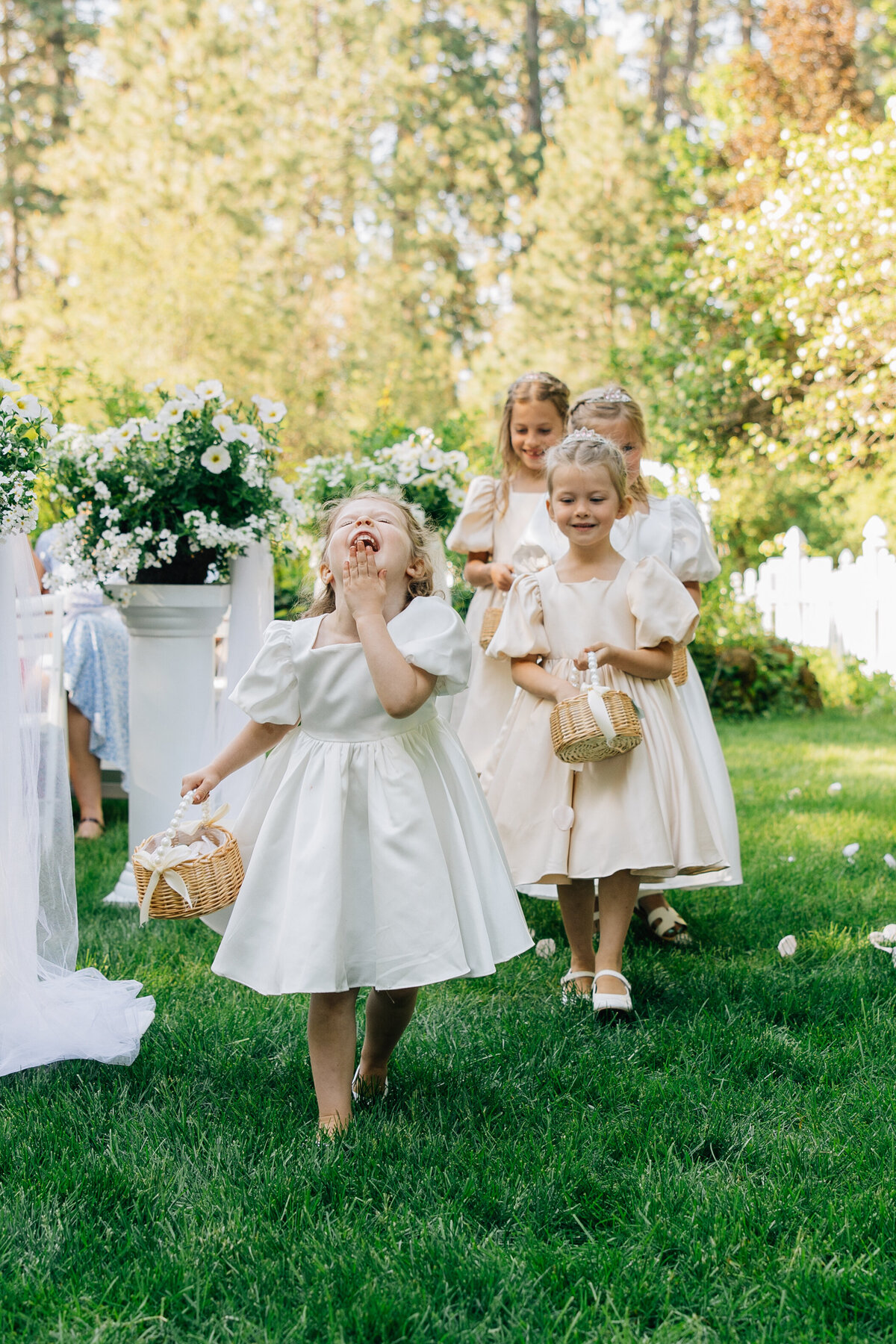 Flower Girls in satin white and tan dresses giggle and smile as they walk down the aisle of a Spokane wedding ceremony at Belle Gardens
