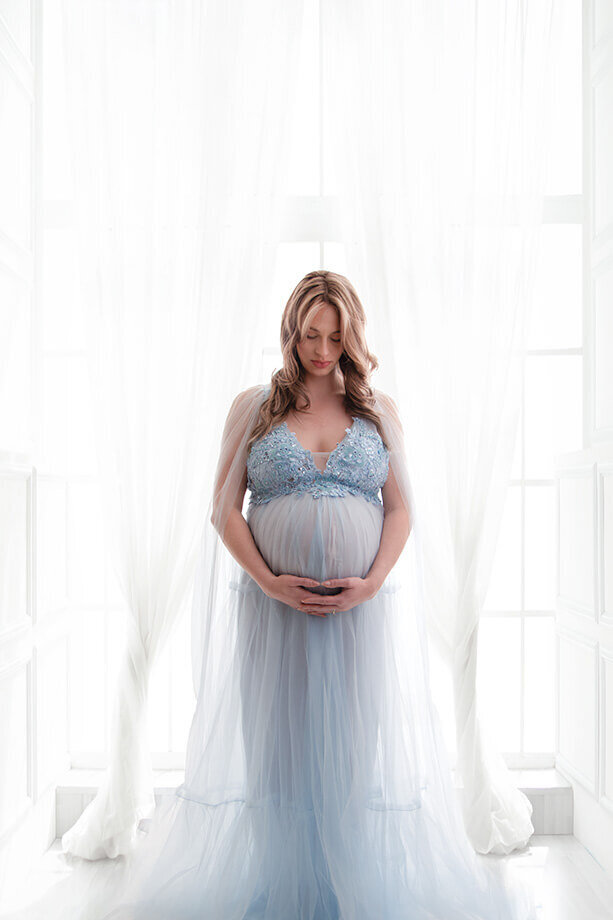 pregnant mother in a soft blue tulle dress in front of glowing chiffon curtains