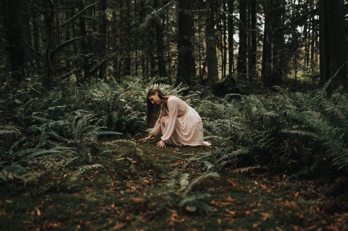 Woman in long, flowy dress sits on lush forest floor and touches the soil surrounded by ferns.
