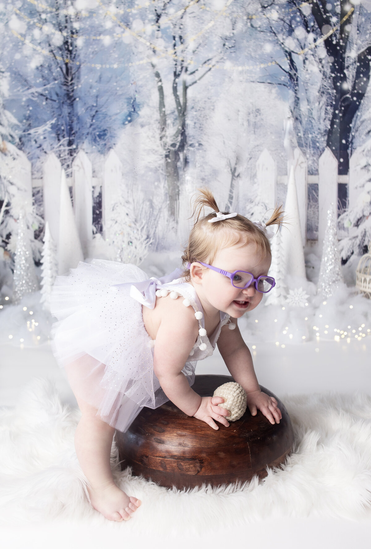 Little girl standing with wood bowl, wearing purple glasses during first birthday cake smash photoshoot in Franklin Tennessee Photography studio. With Franklin Tennessee Family photographer