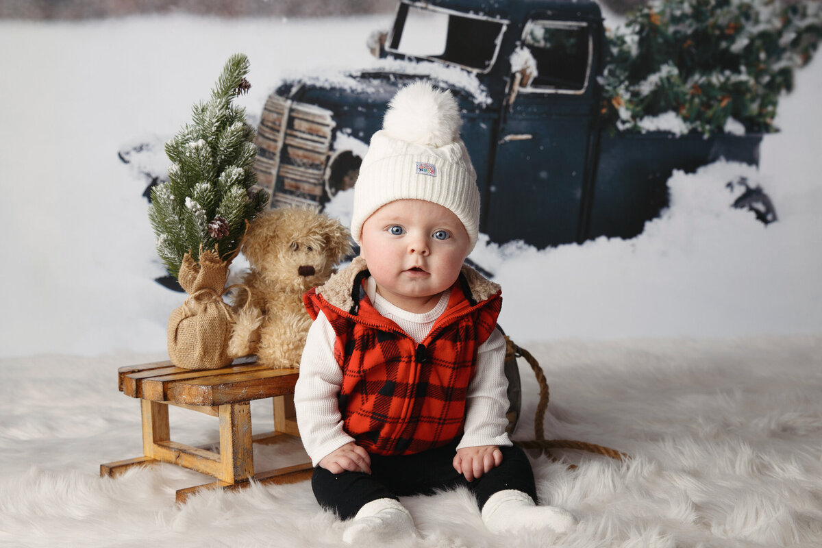 six month milestone portrait of a baby wearing a plaid vest and knit cap sitting in front of a slead and a holiday background
