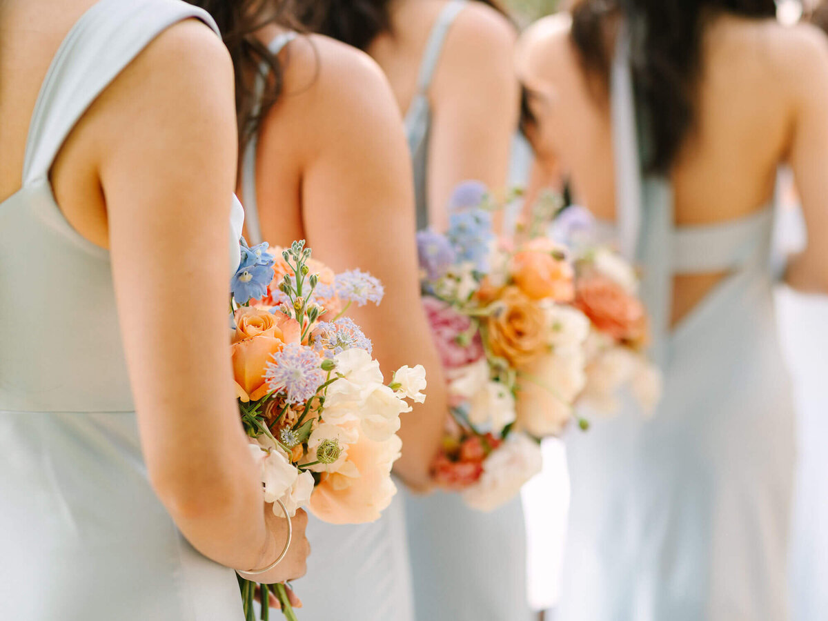 Bridesmaids holding colorful bouquets of orange, pink, and blue flowers, wearing pale blue dresses. The scene is elegant and joyful.