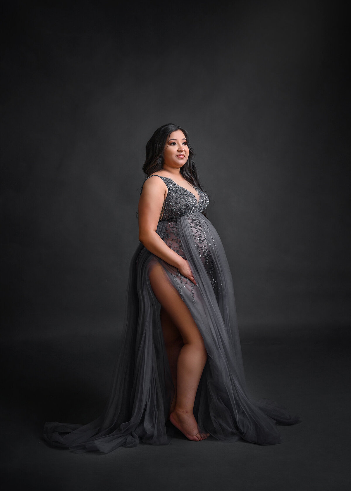 Pregnant woman looking away in a beautiful but simple gray and silver gown with split, in a studio setting for her Phoenix maternity session