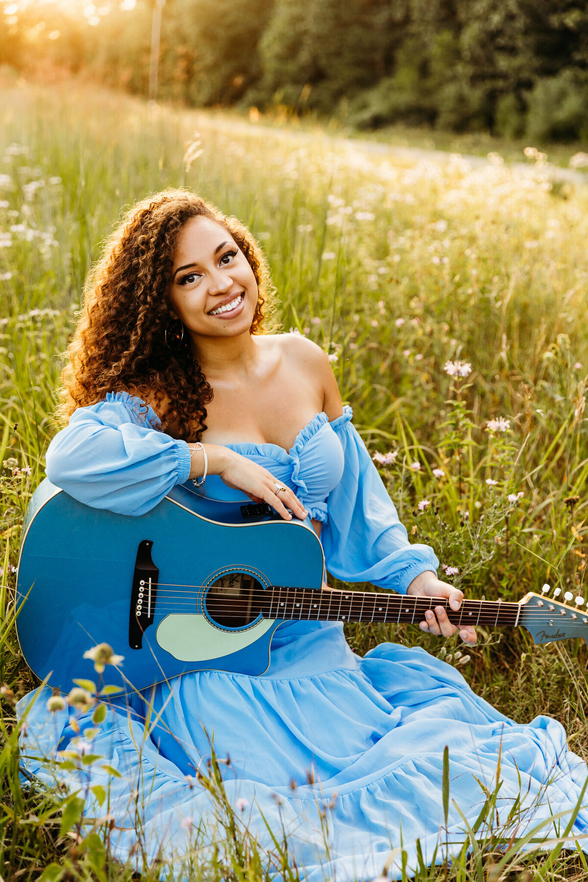 high school senior holding her blue guitar while sitting in a grassy field at sunset