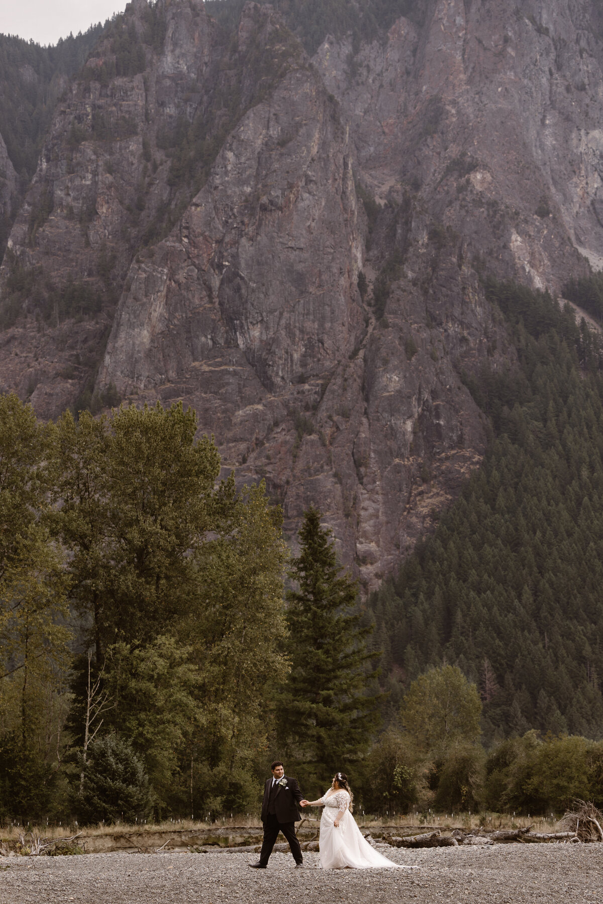 Bride and groom walk hand in hand across a rocky riverbed with towering cliffs and pine-covered mountains behind them, capturing an intimate elopement in the Cascade Range.