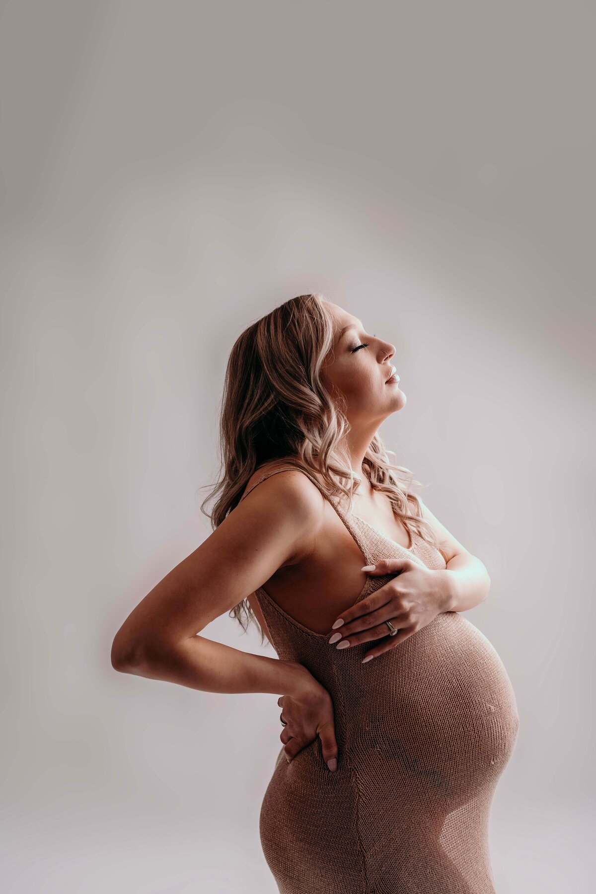 An excited expecting mom poses for a photo showcasing her twin bump. She is wearing a nude sheer dress, with her hand over her chest for modesty. Her other hand rests on her lower nack and she lifts her face upward, toward the light. The photo has nice shadows to create an emotional feeling. Mom looks confident and radiant.