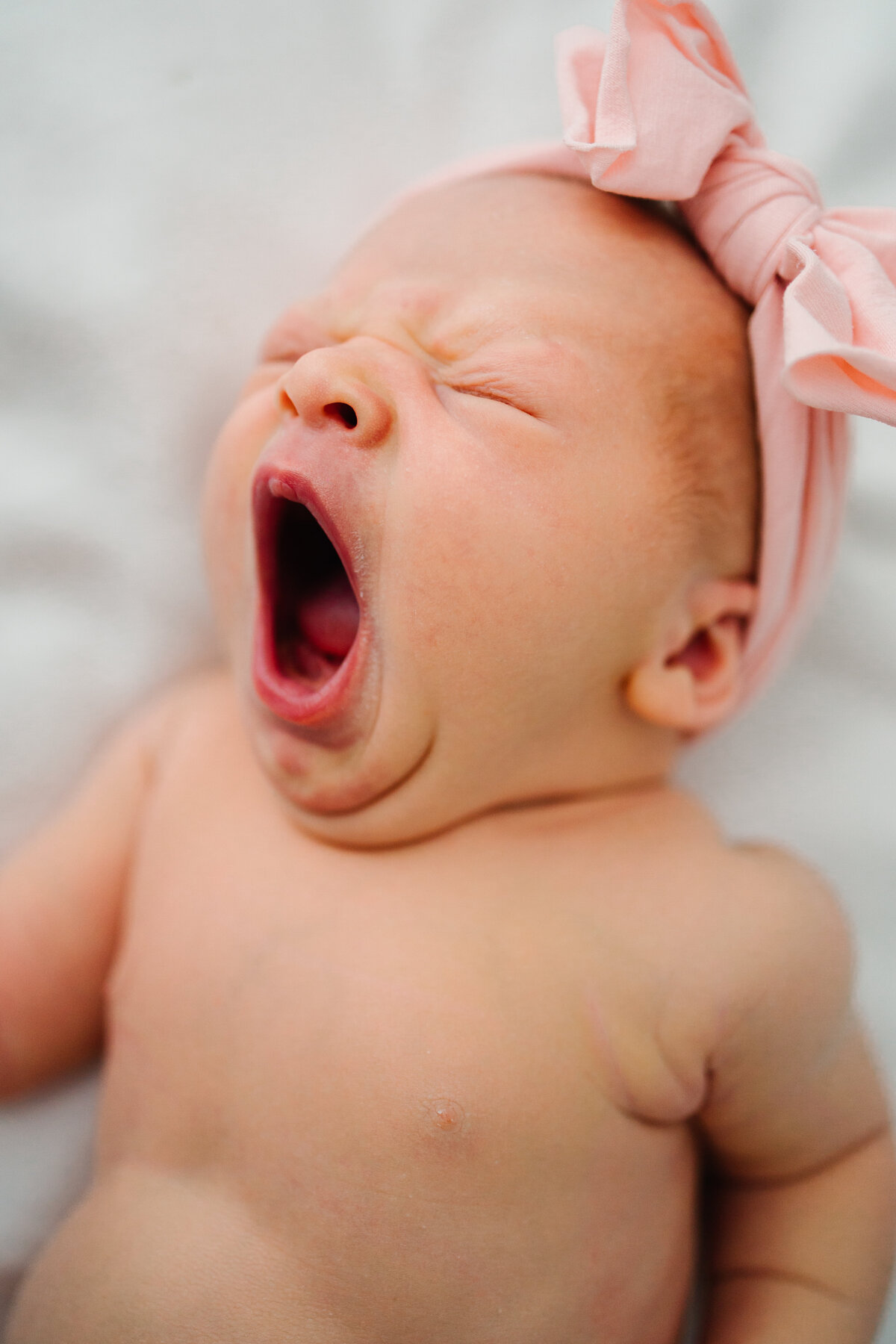 Newborn baby yawning with a baby pink headband on a white blanket