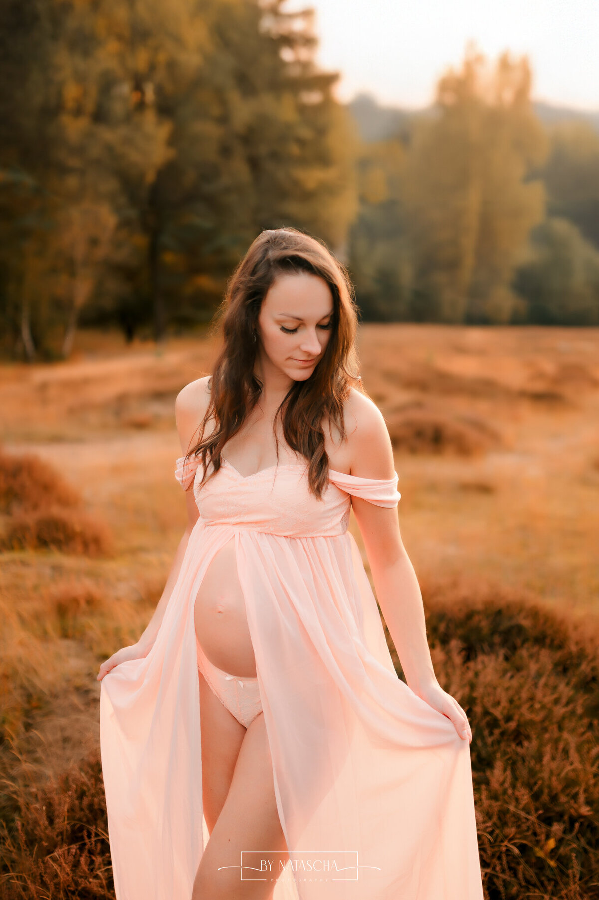Een zwangere vrouw die haar jurk gedeeltelijk openhoudt en haar zwangere buik toont in de heide van Herperduin