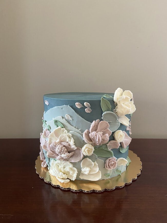 Photo shows a wedding cutting cake with dusty blue frosting. There's partial layers of lighter shades of the same blue around one side, and across the front are pallet knife 3D flowers in mauve and white, with muted green leaves.