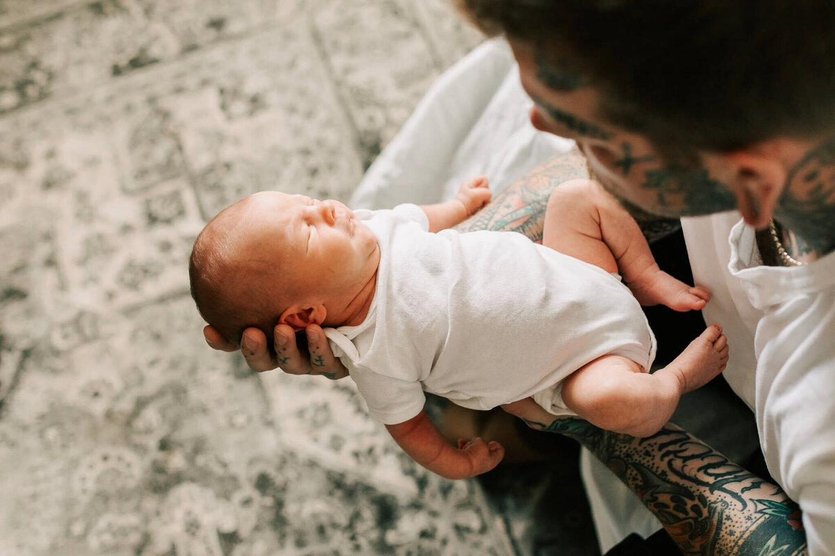 A newborn baby lays in his fathers arms wearing a white onesie