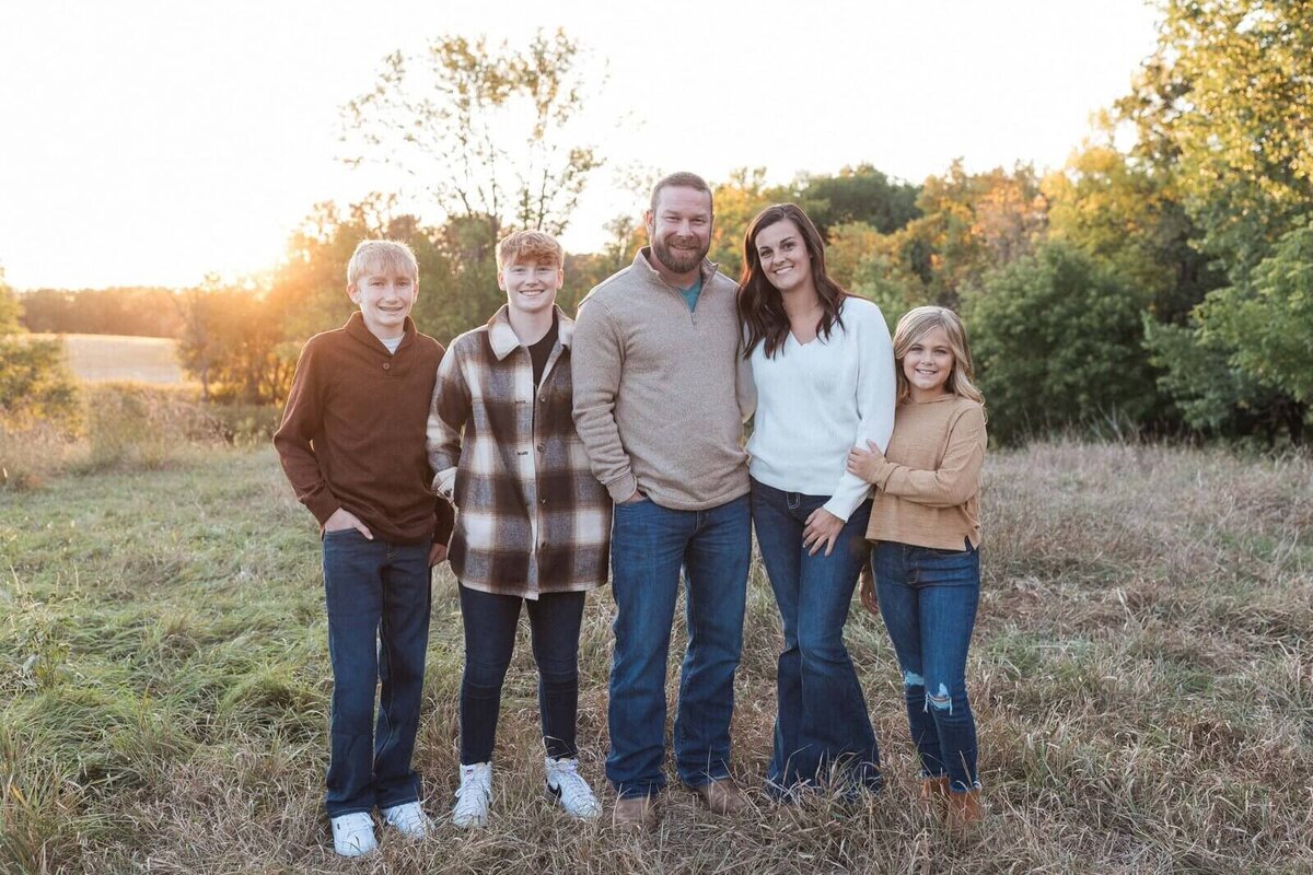 A family of five walks arm in arm through a grassy field, sharing laughs and enjoying a beautiful day.