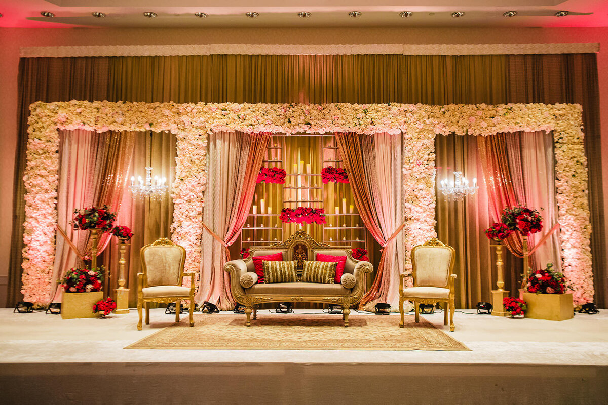 A lavishly decorated wedding stage with a vintage sofa, armchairs, and floral backdrops in shades of pink and gold.