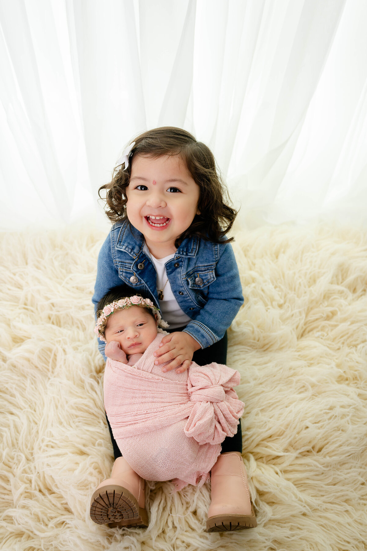 Older child is gently carrying a swaddled baby on her legs with a huge smile on her face. 