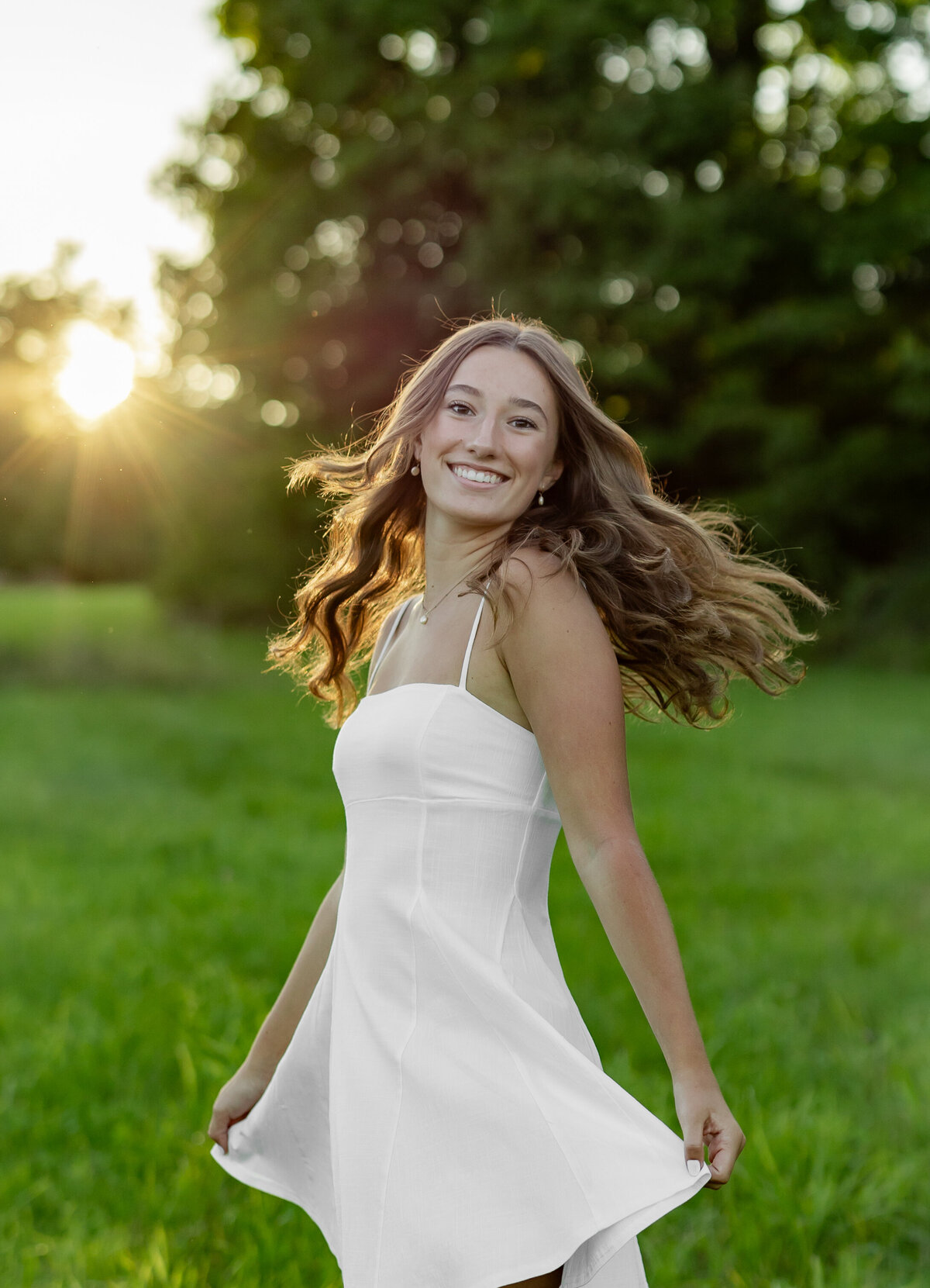 A young woman with long hair in a white dress smiles while twirling in a grassy field. As the sun sets behind her, casting a warm glow, it's a moment perfectly captured by a talented New England Portrait Photographer.