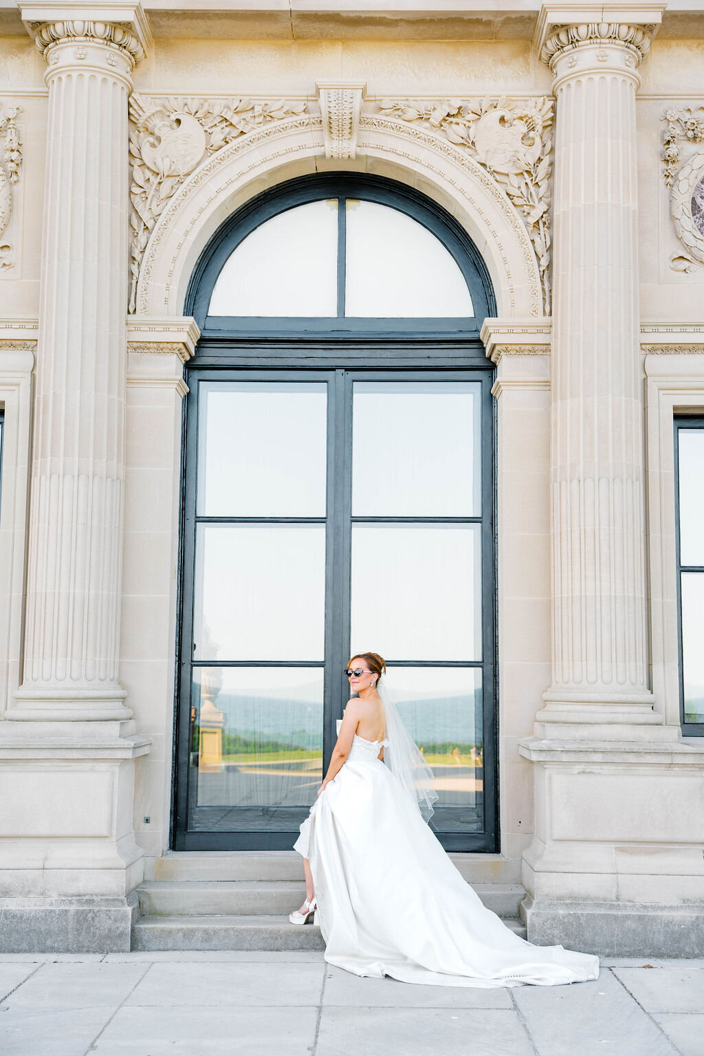 Rhode Island Elopement | Bride in a strapless gown and veil stands confidently before a grand arched window, with elegant columns and stonework.