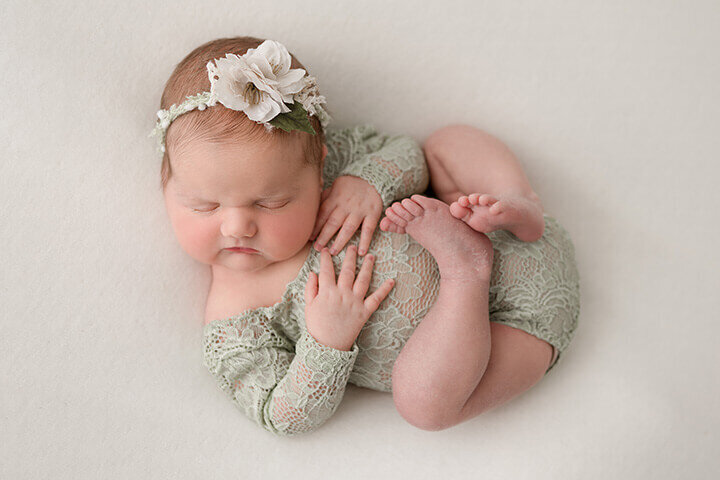 Newborn girl rests on a cream backdrop wearing a sage lace romper and floral headband.