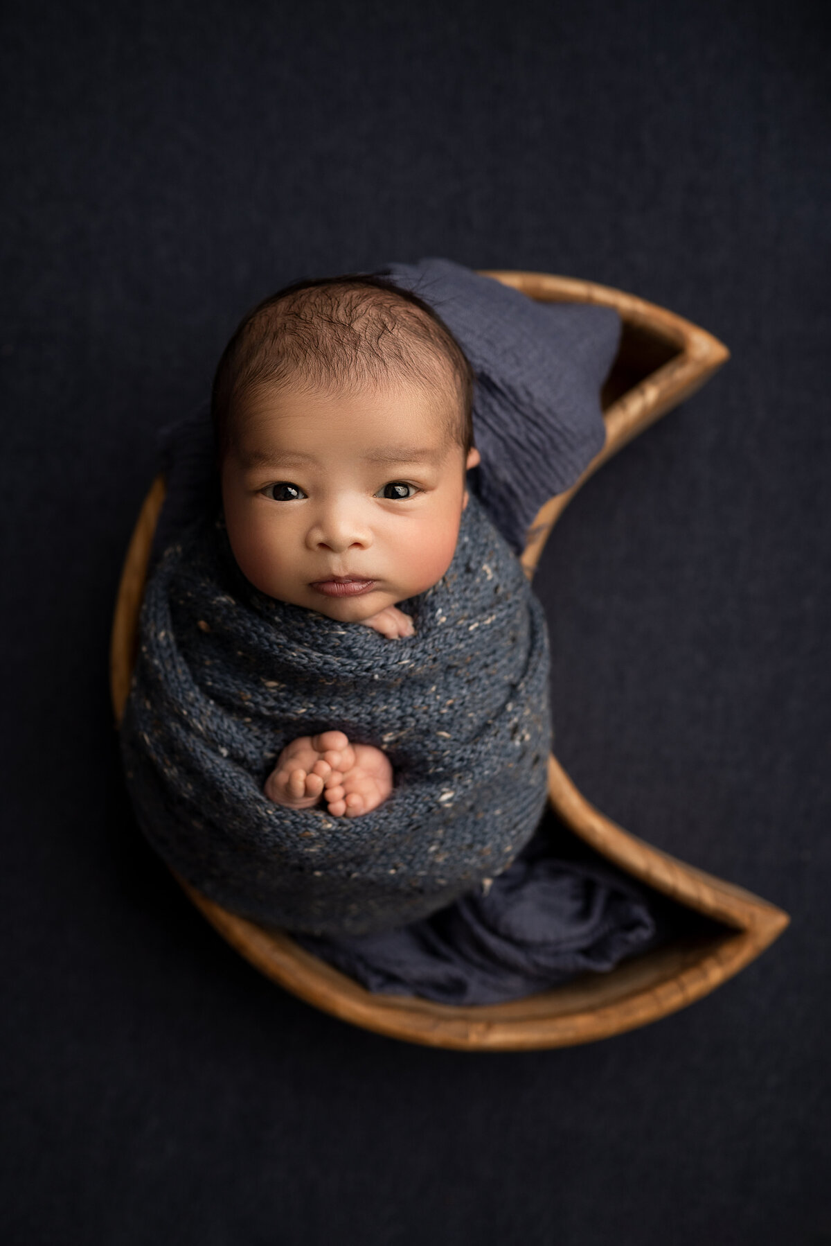 Katie Marshall, New Jersey's best newborn photographer captures a baby boy's fine art newborn photoshoot. Baby is swaddled in a deep blue-grey marled swaddle with his fingers and toes peeking out. He is looking directly at the camera and laying in a moon-shaped bowl. Aerial Image.