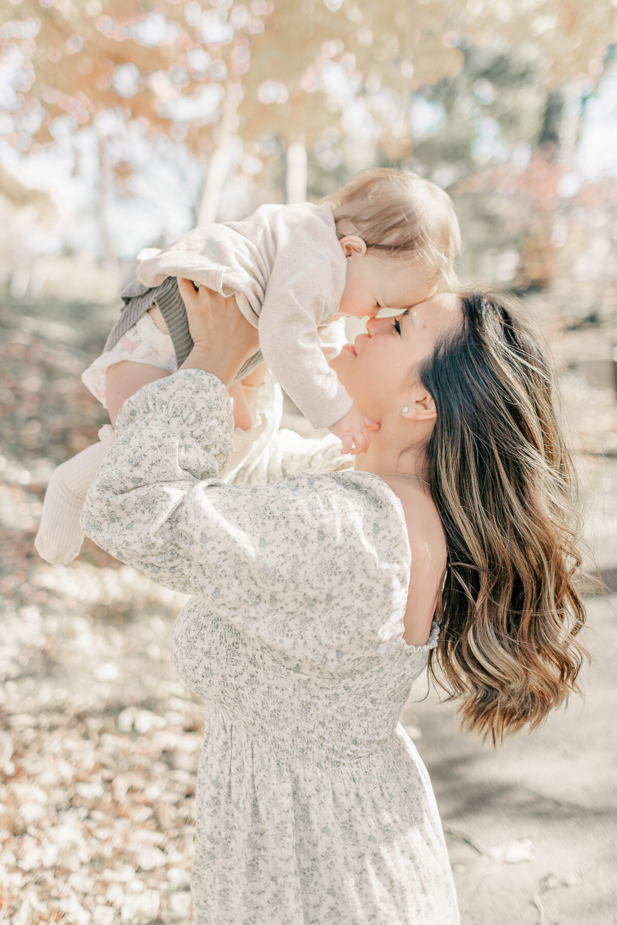 Mother lifting her baby girl in the air while surrounded by fall leaves during a Kansas City first birthday milestone session.