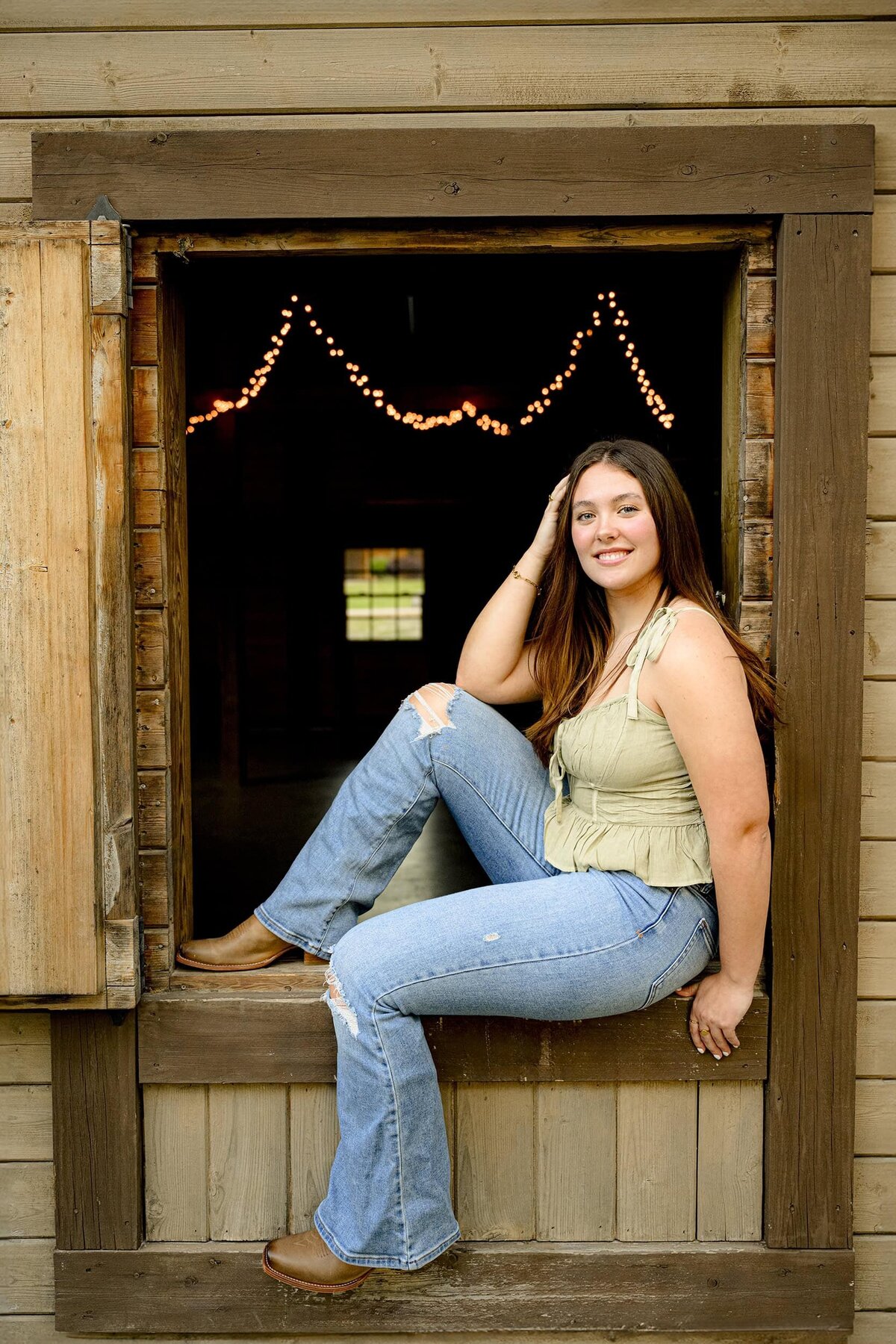 charlotte area senior sits in barn window wearing jeans and a green top for her senior portraits