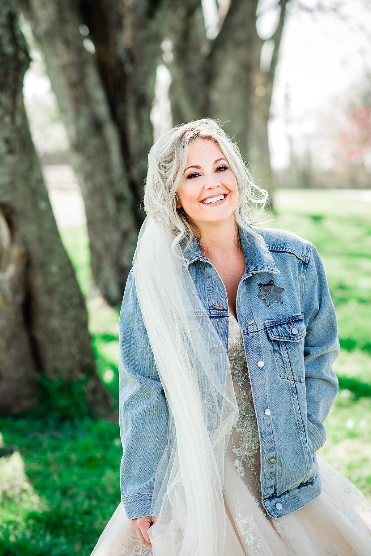The bride, wearing a long sheer veil and denim jacket smiles up at the camera as she stands beneath a tree.