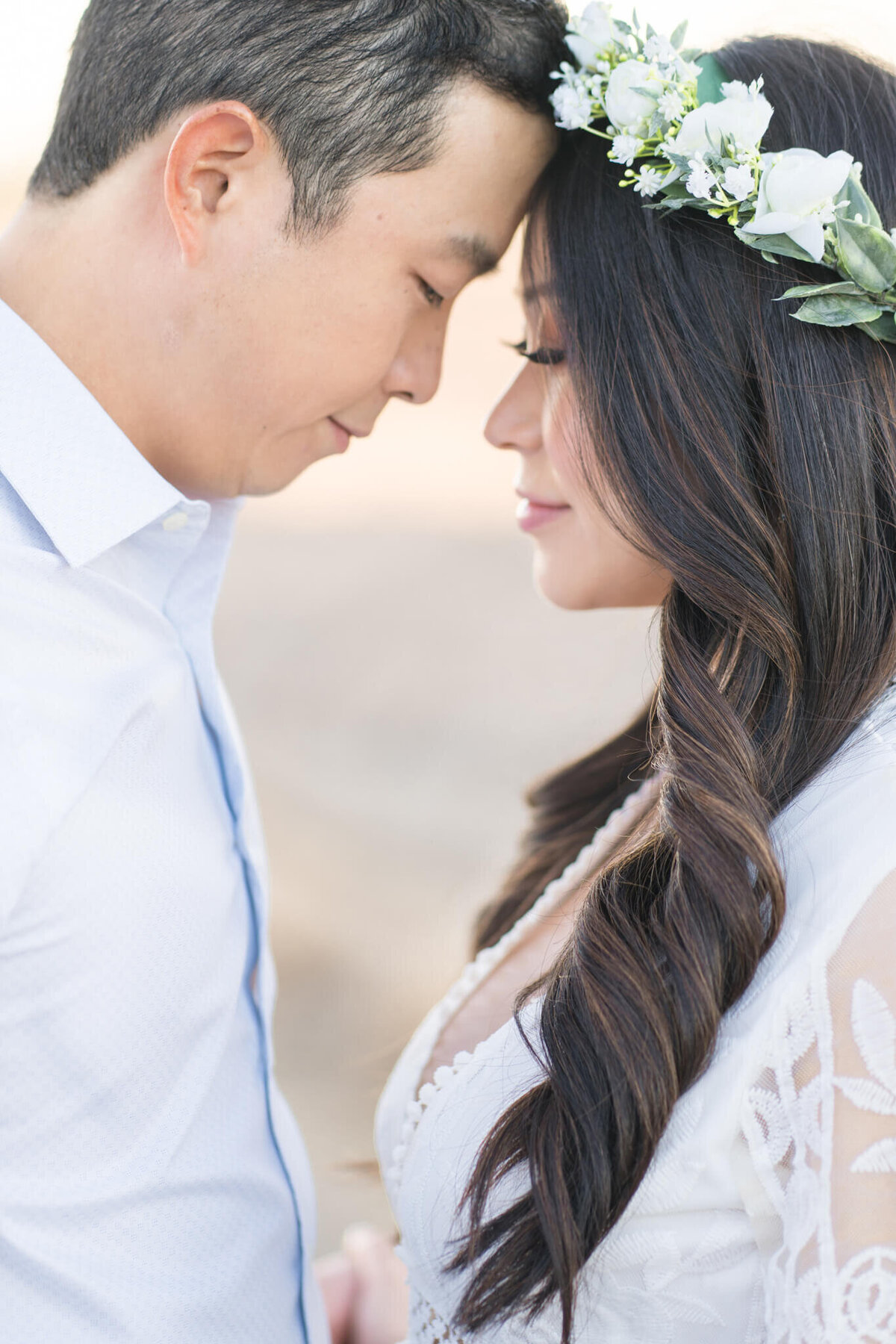 Asian couple touching foreheads and holding hands. White dress and flower wreath. By Orange County Maternity Photographer Melliemade Photography