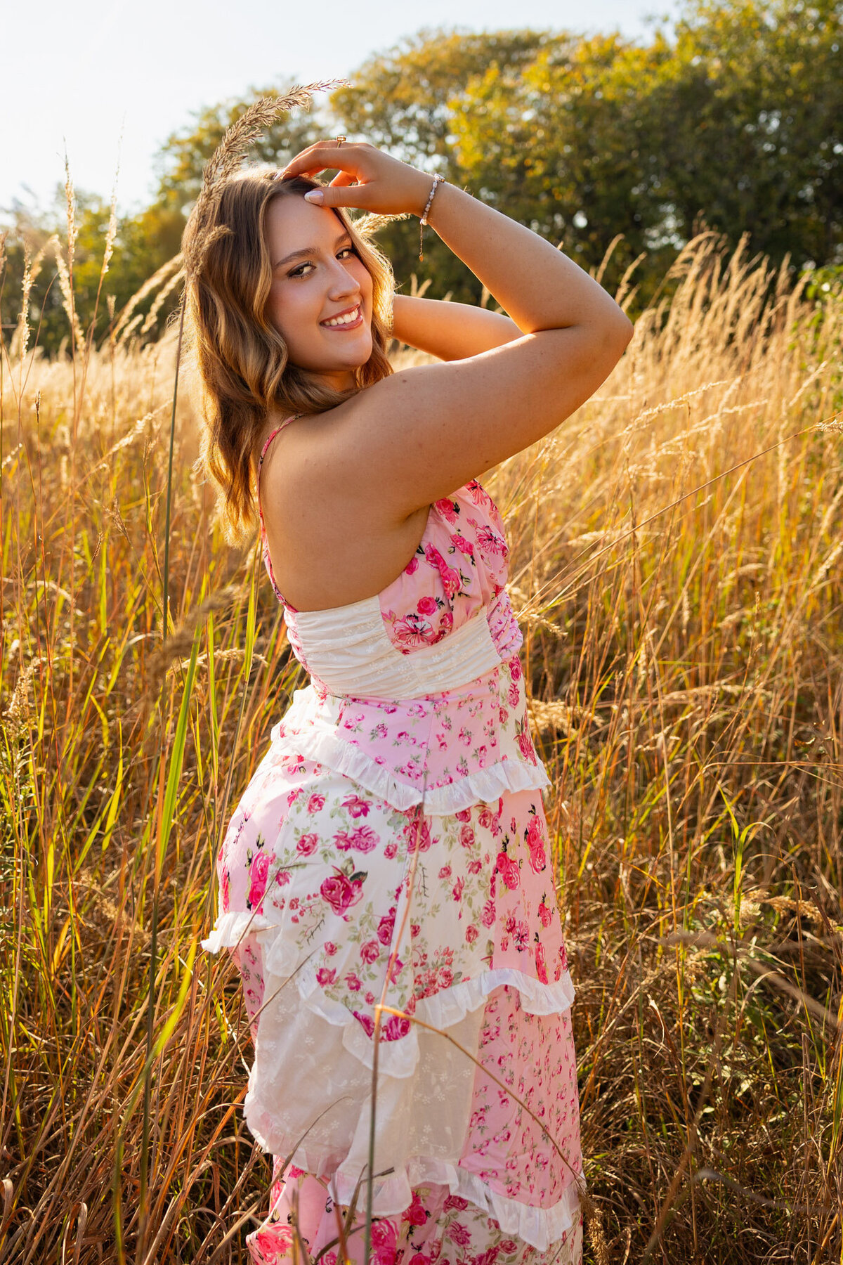 Senior girl posing in tall grass with hand on hat at sunset in Lawrence KS