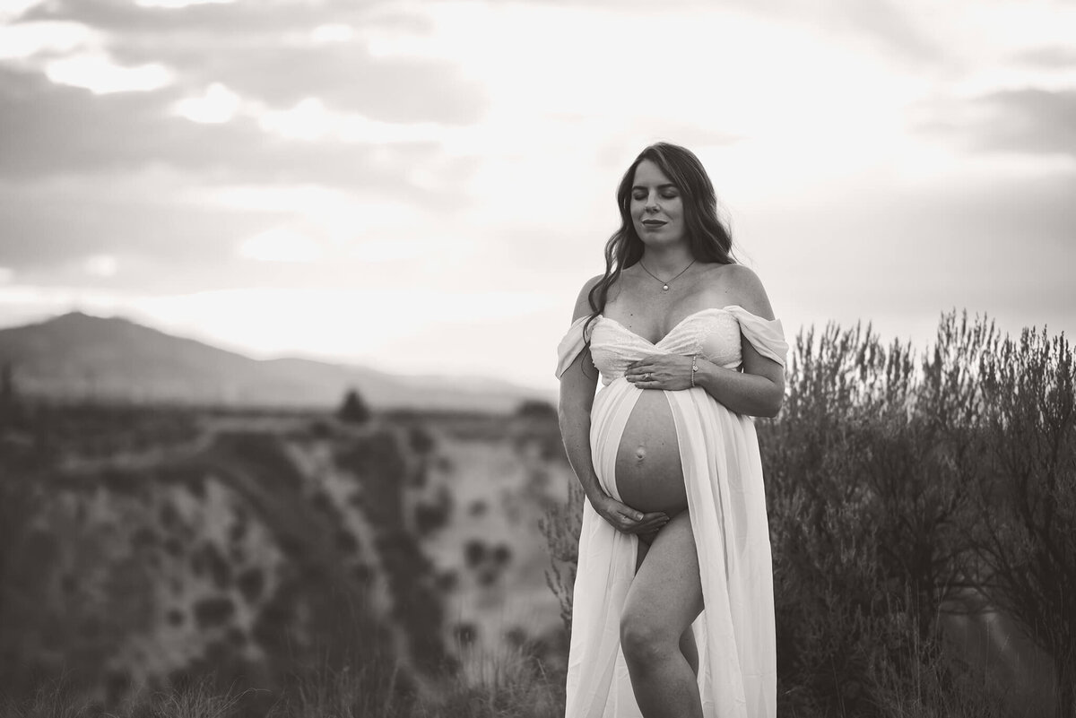 Brittany Danielle takes a black and white images of a pregnant mother wearing a white flowy dress while holding her belly. Taken in Kamloops