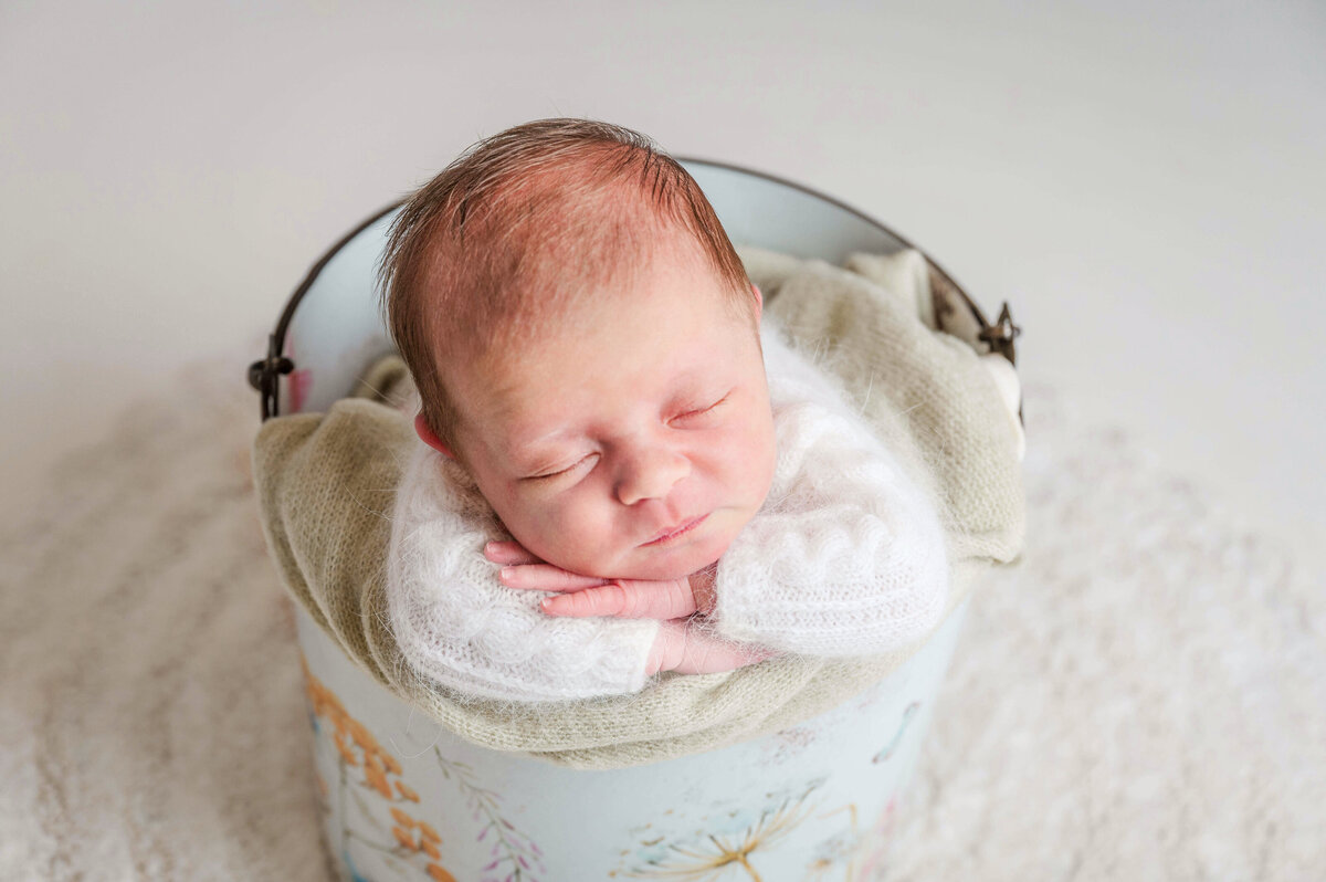 Precious newborn in fuzzy sweater laying with her chin on her arms in a cream bucket with painted flowers.