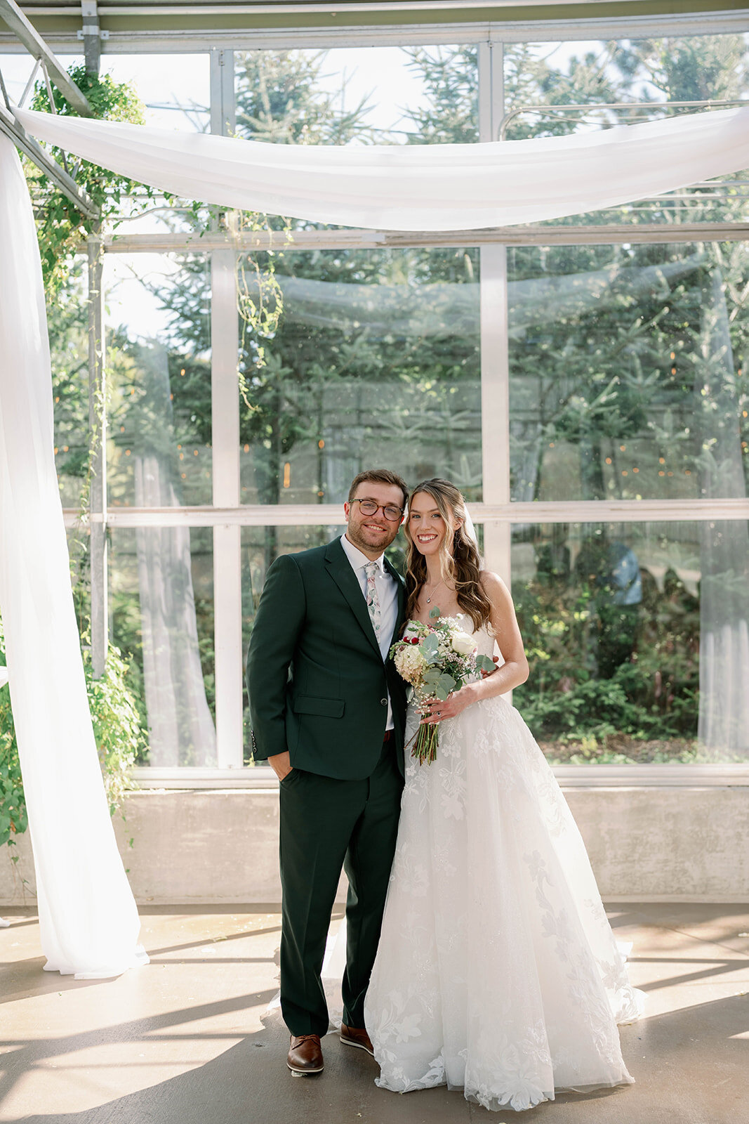Traditional portrait of the bride and groom smiling at the camera inside the greenhouse at The Ivy House wedding venue.