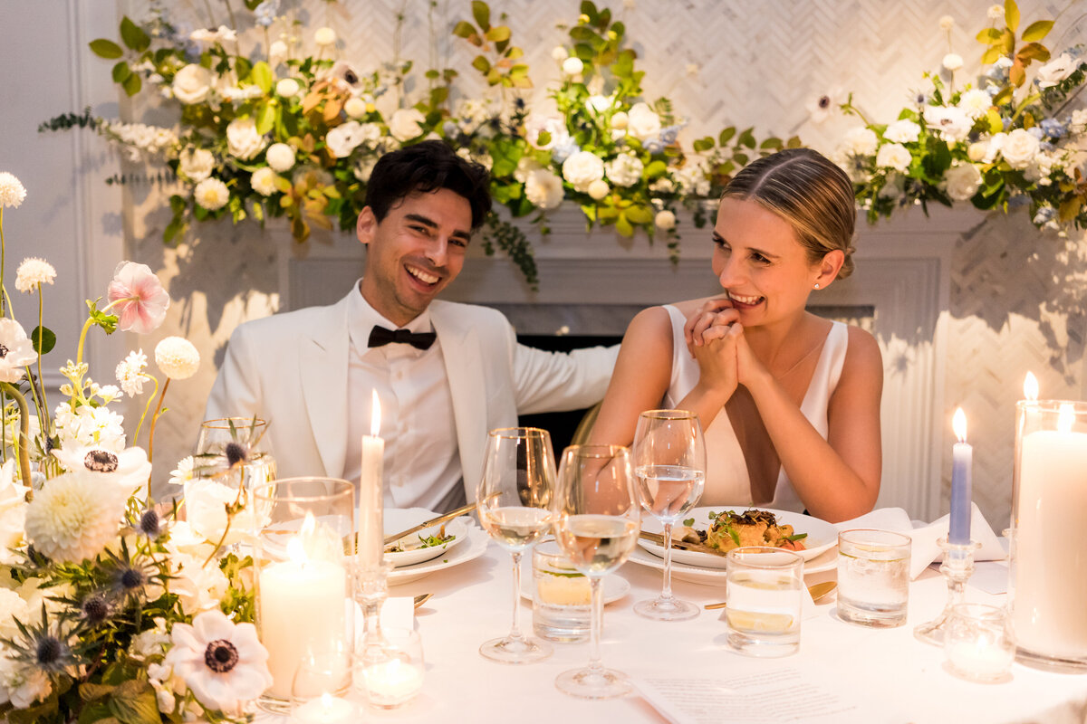 A bride and groom smile at a table at the Atwater Club decorated with white flowers and candles.