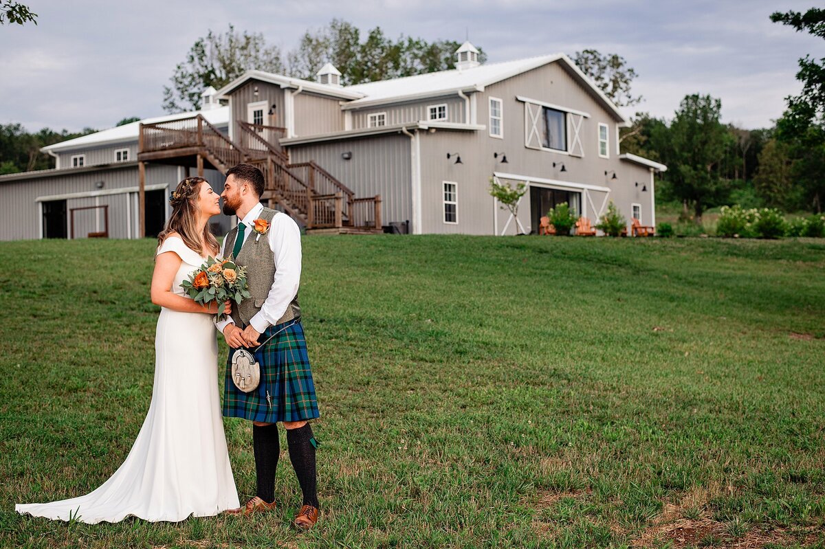 The bride, wearing a long off the shoulder wedding dress with a flowing train holds a bouquet of orange and yellow roses with eucalyptus and greenery as she leans in to kiss the groom. The groom is wearing traditional Scottish attire. A white shirt with a gray vest with a blue and green plaid tartan kilt and ivory sporran with tall wool socks and brown shoes. Behind them up on a hill is a large gray barn. Lush green rolling hills and tall cedar trees.