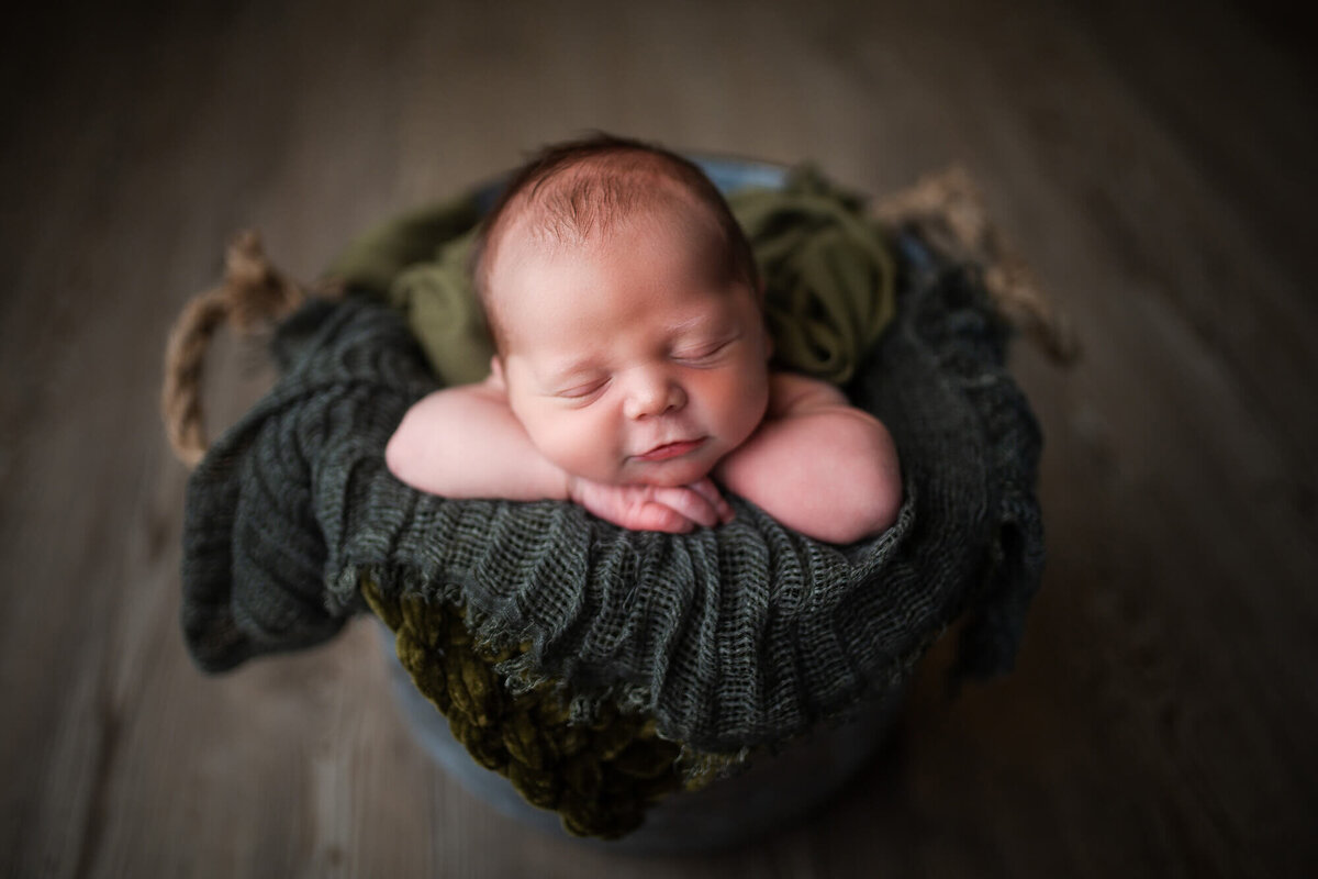 In home newborn portrait. Baby boy is smiling in silver bucket laying on rustic natural green tones 