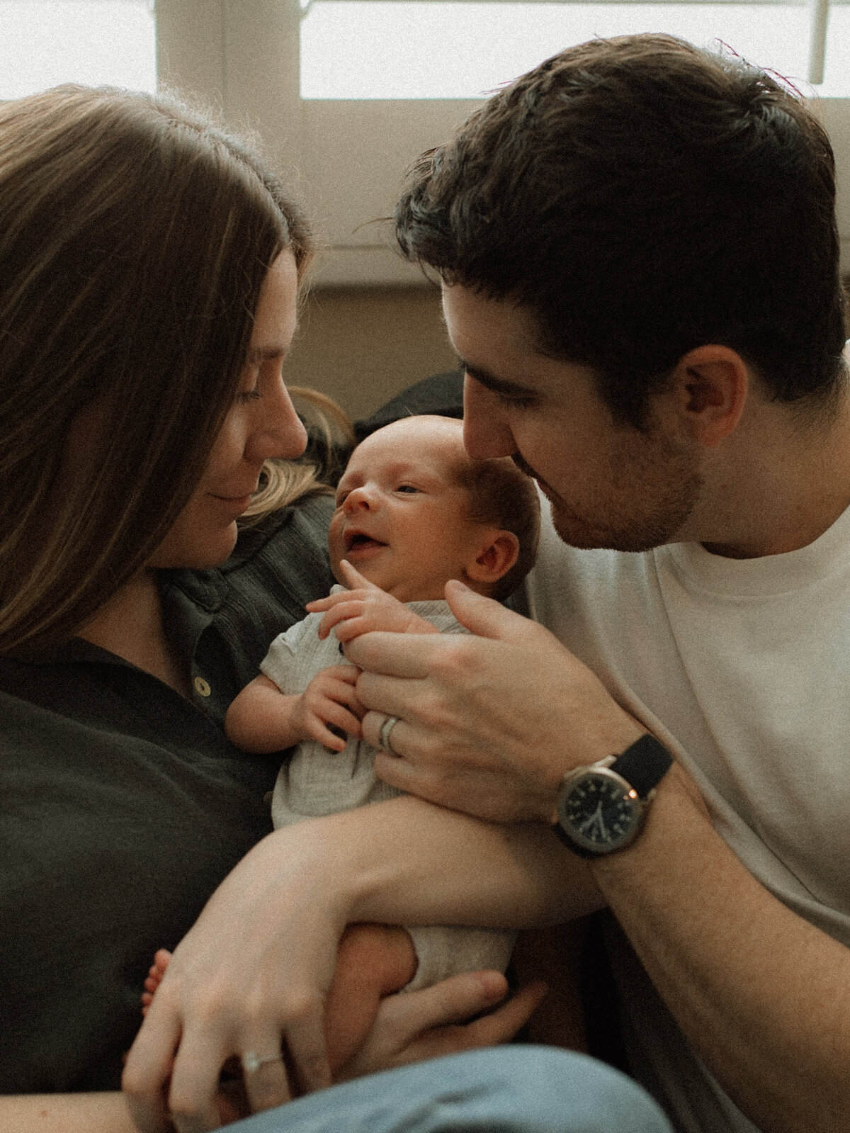 A mother and father smiling at their newborn as they cradle the baby between them, lit by soft natural window light.