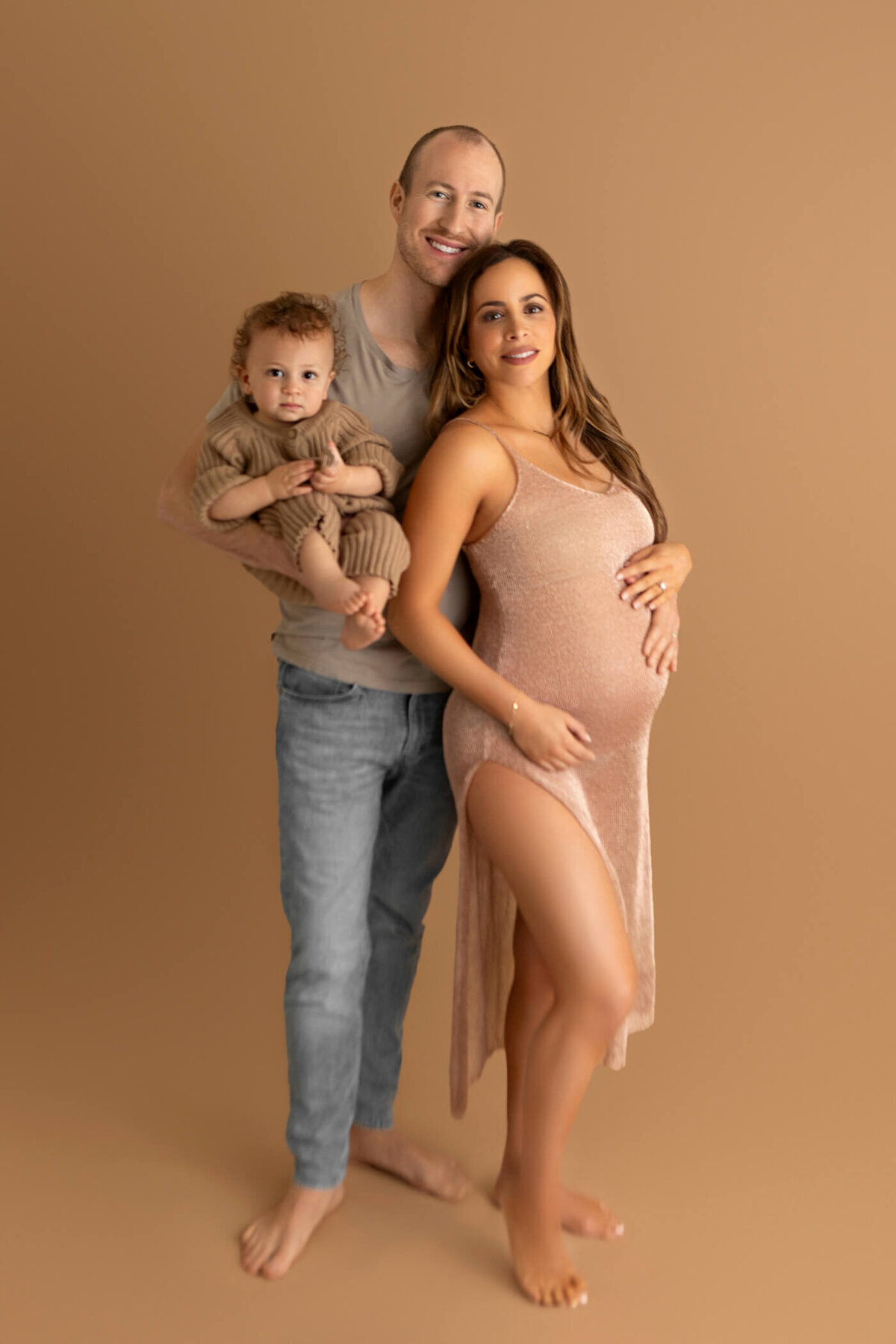 A happy family stands together against a warm beige background. The pregnant mother is wearing a shimmering, sleeveless gold dress that accentuates her baby bump. She stands next to her partner, who is holding their young child. The partner is dressed casually in a beige T-shirt and jeans, while the child is wearing a cozy, brown outfit. All three are barefoot and smiling, creating a warm and loving family portrait.