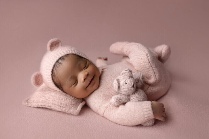 A newborn baby smiles in her sleep in a knit onesie with a bear hood and matching knit stuffed bear in a New Orleans Newborn Photography studio