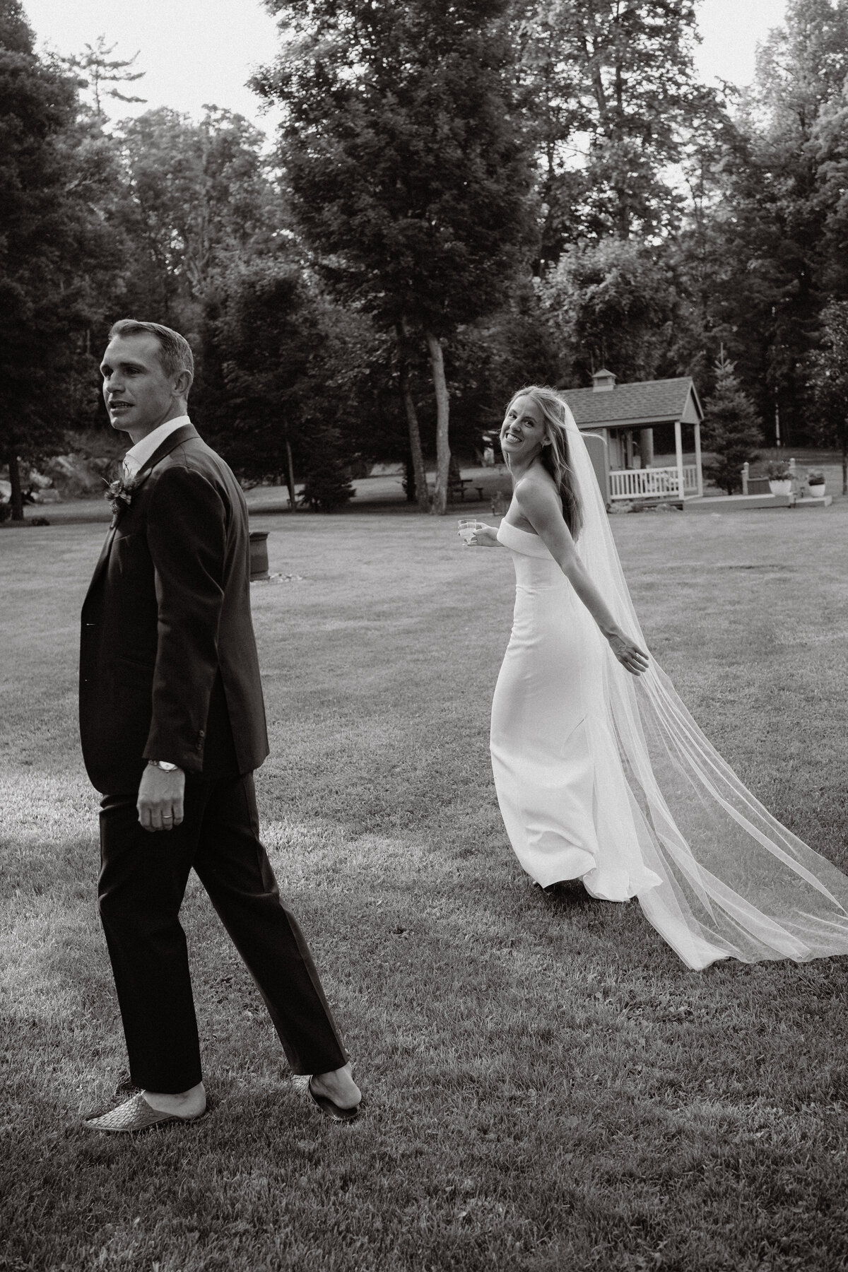 the newly married couple walks through the cottage property with champagne looking back and smiling