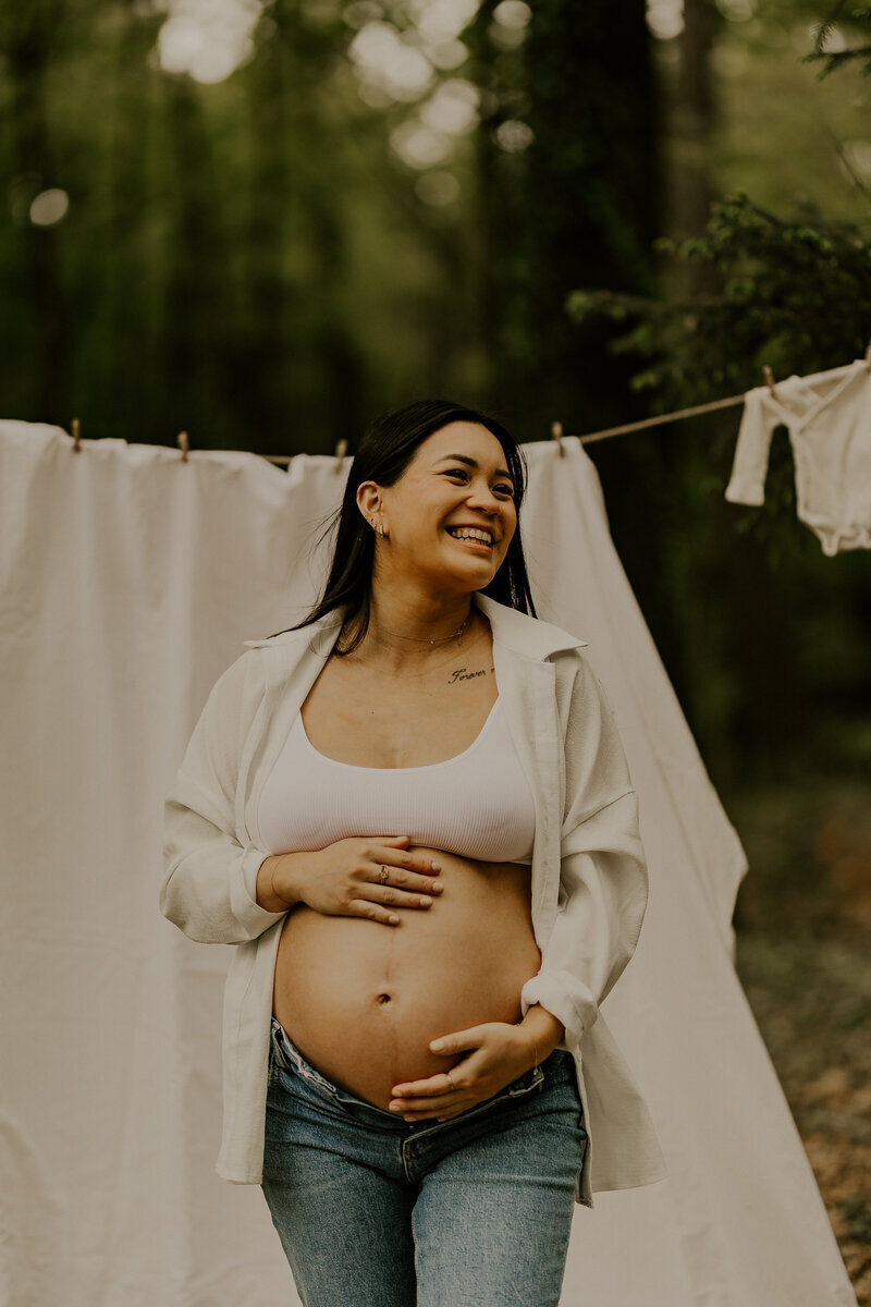 Femme enceinte, souriante, en jean et cehmise blanche, ventre découvert pose en forêt devant du linge étendu sur une corde. Séance photo grossesse en vendée.