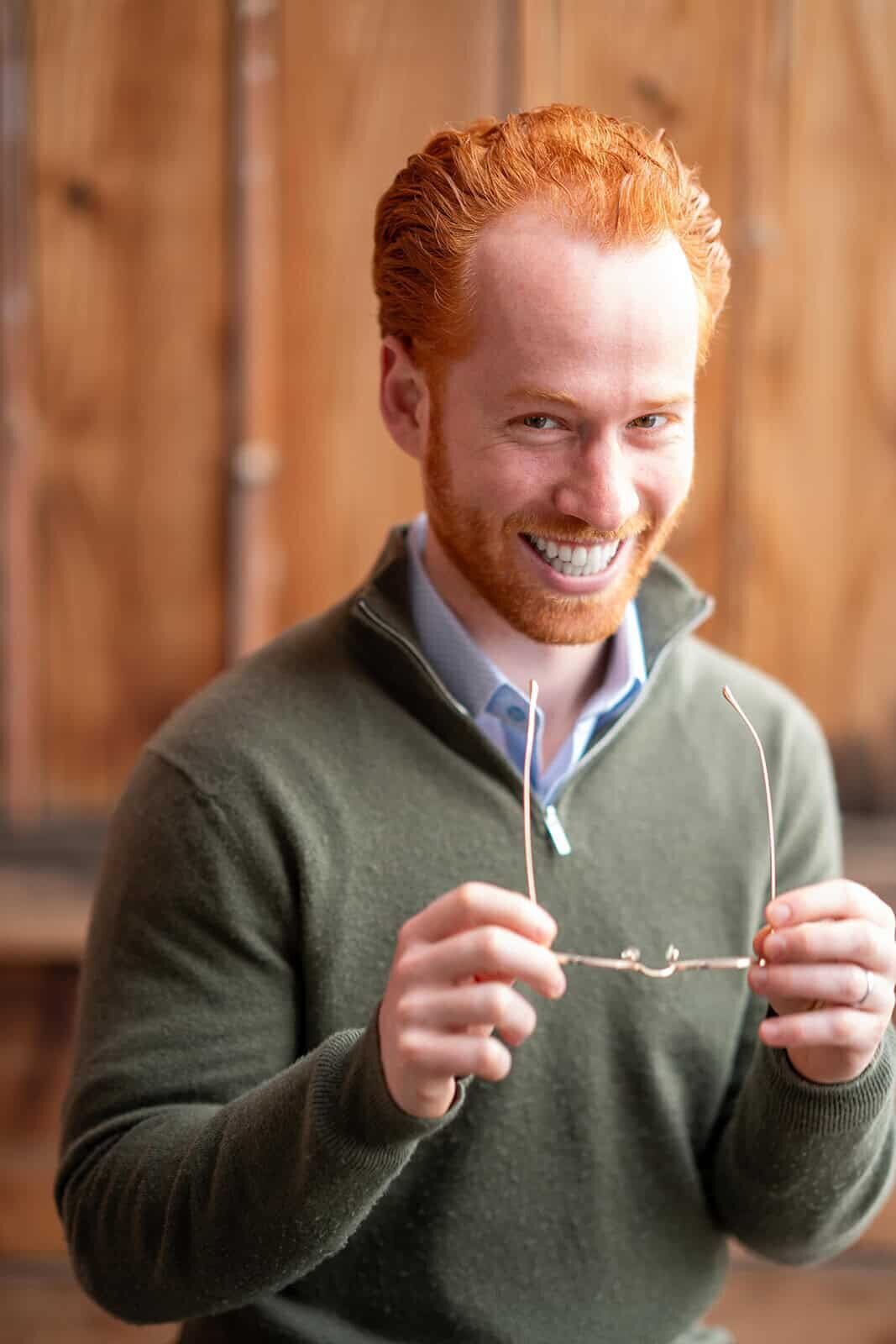 Bearded man wearing a sweater, holding glasses at chest level, and laughing