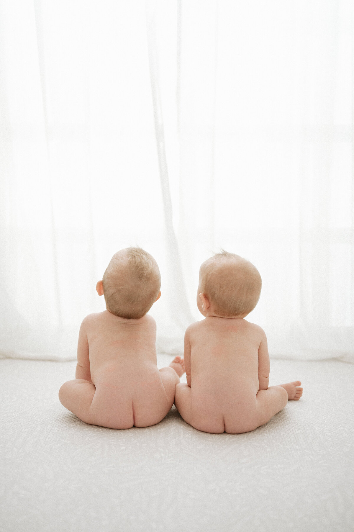 Two naked babies sit side by side looking out a window