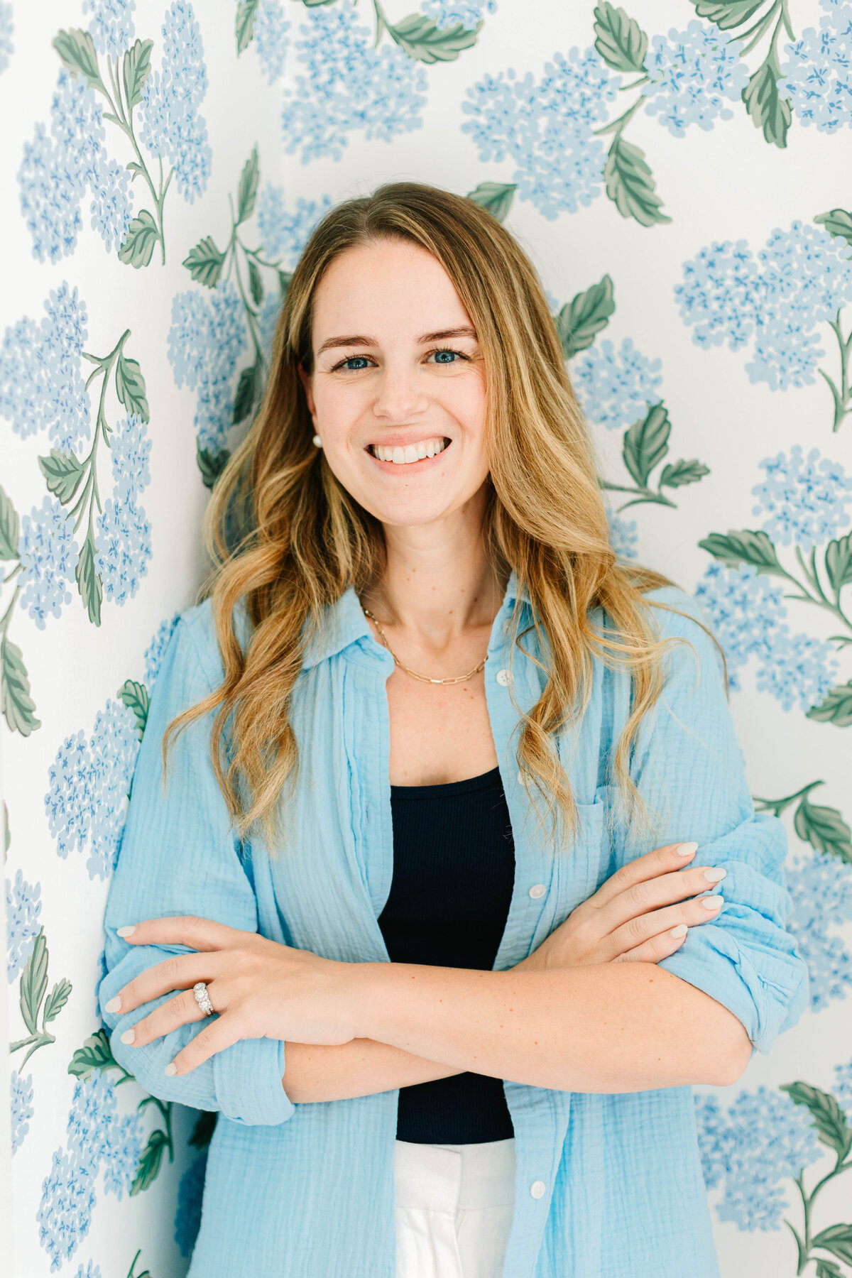 A smiling woman with wavy blonde hair stands with her arms crossed against a floral wallpapered wall featuring blue hydrangeas. She wears a light blue button-up shirt over a navy top, with white pants and a gold necklace.