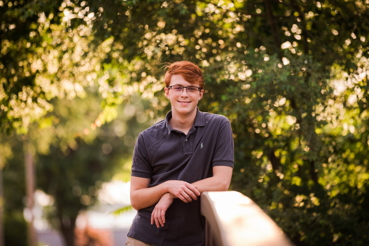 A high schooler young man with red hair and glasses leans sideways on a bridge in front of trees.