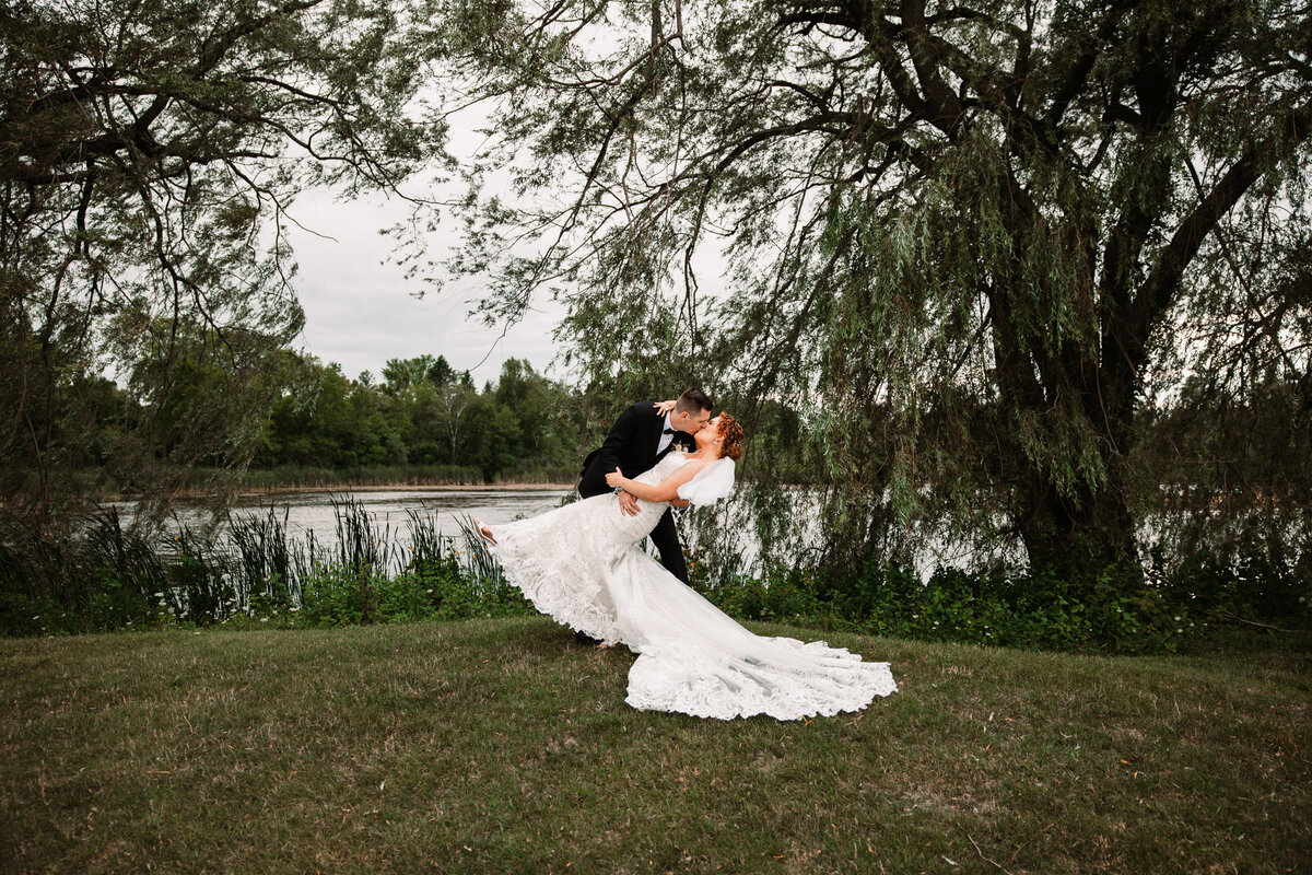 Newlyweds kiss and dip under oak trees by a large pond