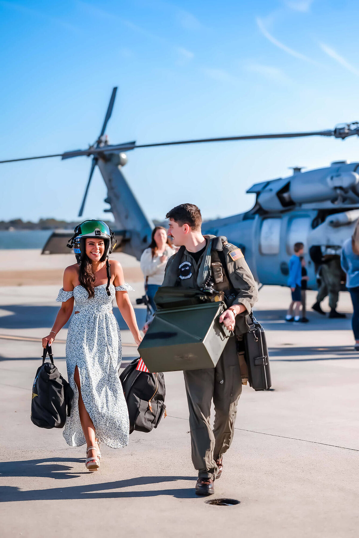 A military spouse wears her husbands flight helmet and carries two of his bags while he looks back at her. Photo taken by Justine Renee Photography during a helicopter homecoming at Norfolk Naval.